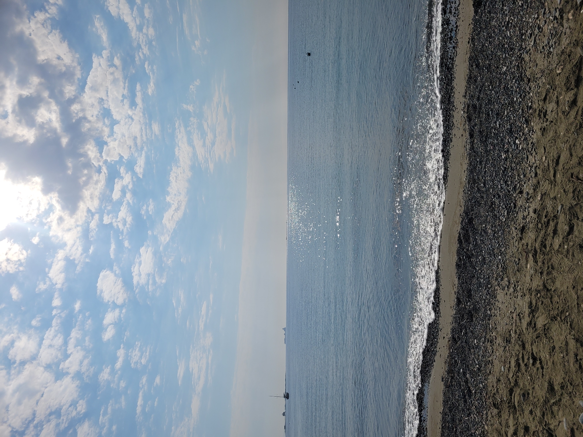 The image depicts a serene coastal scene in Tserkezoi, Cyprus, on a sunny day. The foreground features a sandy beach transitioning to a pebble-strewn shoreline. Gentle waves with white foam are lapping at the edge of the sea. The vast expanse of the calm, blue ocean stretches towards the horizon, reflecting the bright sunlight, creating shimmering patterns on the water's surface. In the distance, several buoys are visible, marking a section of the sea. Further out, a sailboat and a large cargo ship can be seen on the water, adding a touch of maritime activity to the otherwise tranquil landscape. The sky above is a clear blue, dotted with scattered white clouds, with the sun partially obscured by clouds in the upper right corner, casting a bright glow.