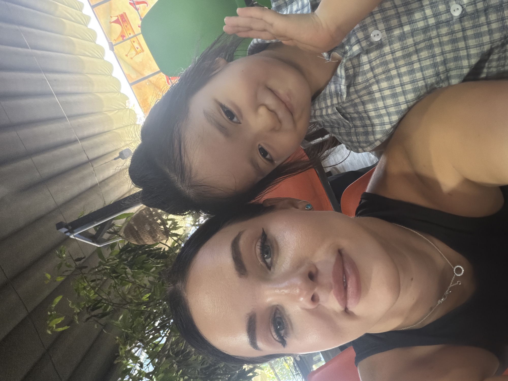 A selfie taken by an adult woman with a young child. The woman has dark hair, light skin, and wears a black top and silver necklace. The child has dark hair and is wearing a light blue and white checkered shirt. They are likely sitting together at a table, as hinted by the orange and green chair backs. The background shows a partially obscured roof structure, foliage, and signs. The location is in the city of Nam Nha Trang Ward, Vietnam.
