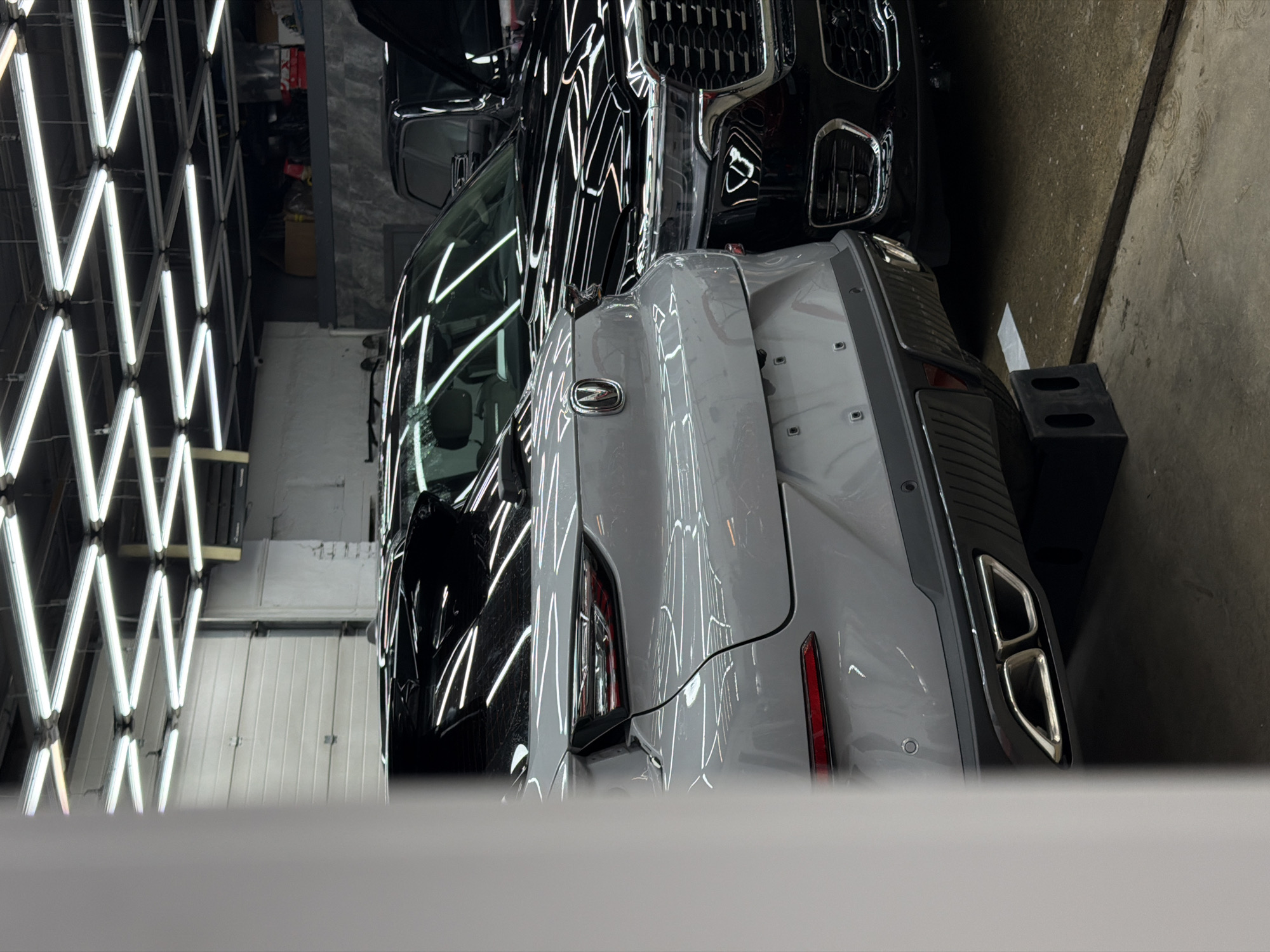 An interior view of a commercial vehicle service or detailing center in Krasnoyarsk, Russia, illuminated by an overhead grid of fluorescent lights. Two SUVs are parked within the bay. In the left foreground, the rear-left quarter of a light grey Changan SUV, identified by its badge and dual rectangular exhaust tips, is visible. Its bodywork is highly reflective, showing sharp reflections of the overhead lighting. The vehicle's rear tires rest on black chocks. To the right and slightly behind the Changan, the front-right quarter of a black SUV, featuring a prominent chrome grille with vertical slats and multi-element headlights, is visible; this vehicle resembles a Jeep Grand Cherokee L. Its black paint also exhibits significant reflections of the lighting. The floor is concrete, displaying wet patches or stains. In the background, a large white sectional garage door is partially visible on the left, and a raised storage platform with various boxes is present on the right. A light-colored vertical architectural column or wall obstructs a portion of the left foreground. No individuals are present in the scene.