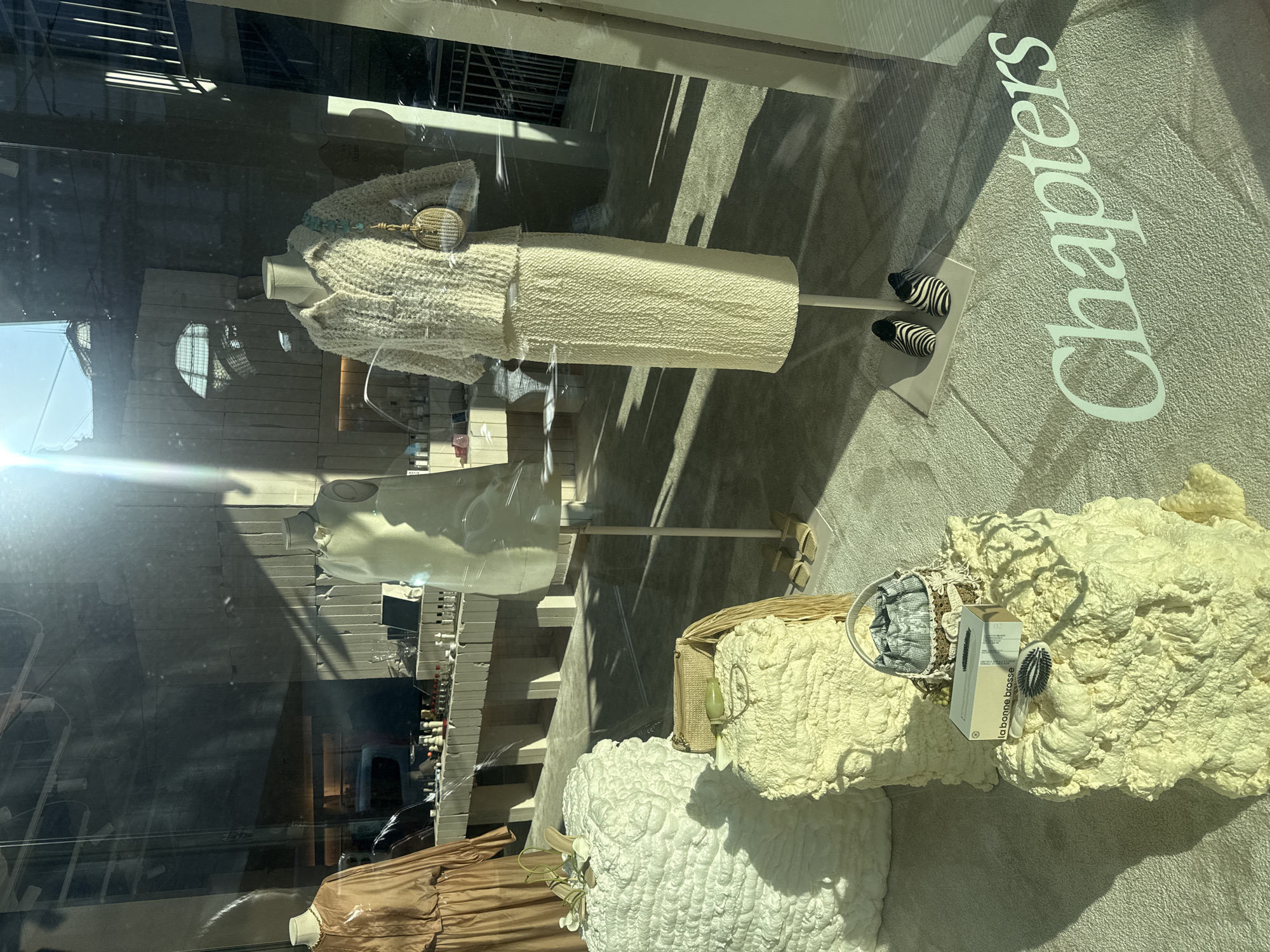 The image depicts a shop window display in Milan, Italy. The store, identified by the "Chapters" signage on the floor, showcases a selection of clothing and accessories. Mannequins are dressed in textured, cream-colored garments. Other items are displayed on similarly textured platforms. The window reflection shows the exterior of the building across the street.