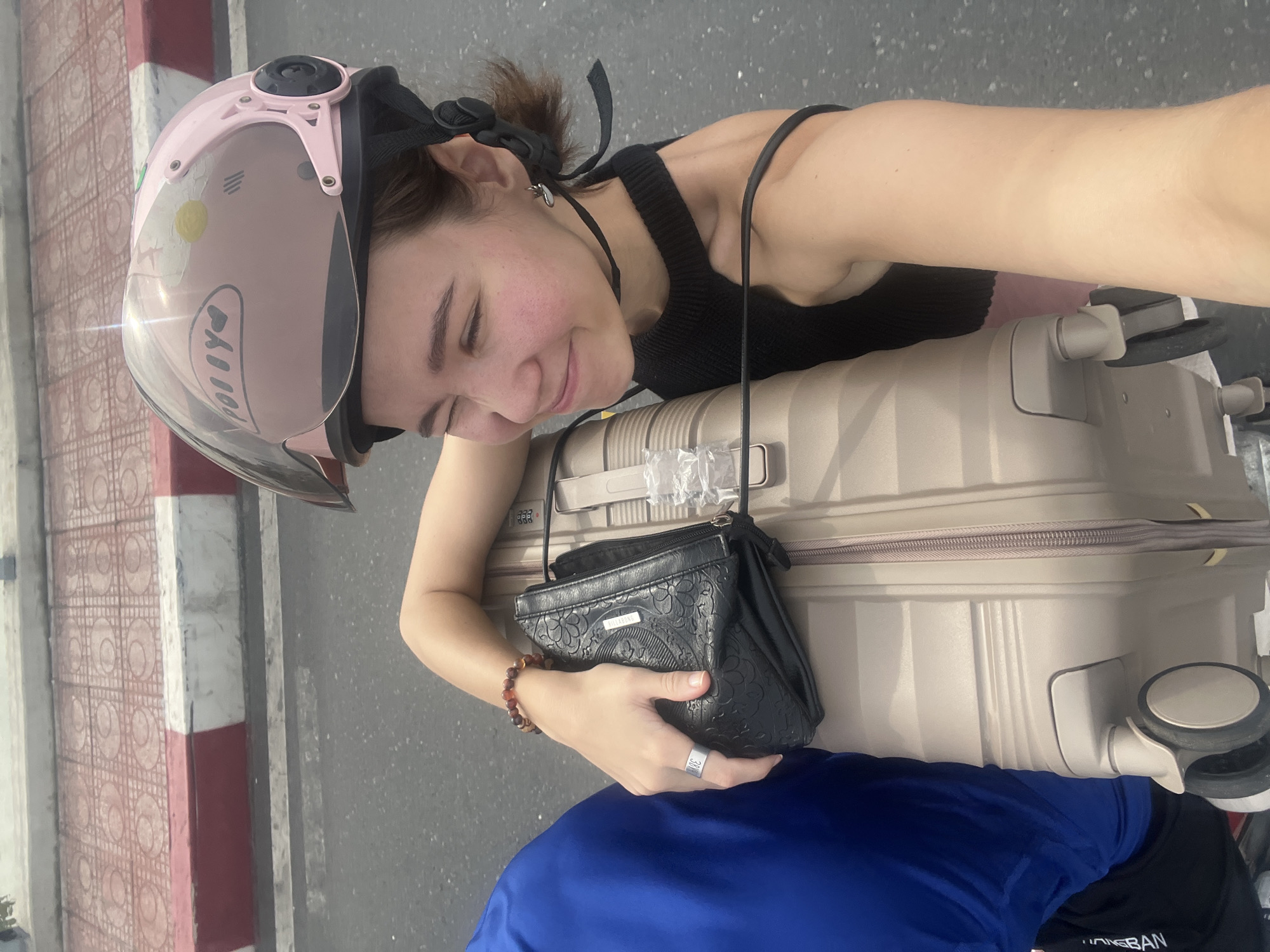 This image captures a young woman traveling or awaiting transport on what appears to be a scooter or motorcycle in an outdoor urban setting in Bac Nha Trang Ward, Vietnam. The scene is illuminated by bright daylight, suggesting a clear and sunny day.
The primary subject is a young woman, seen from a slightly high angle. She wears a pink open-face helmet with a clear visor, which has a yellow daisy sticker and the reversed text "POLIYO" visible from a reflection. Her eyes are closed, and she has a gentle, contented smile on her face, with a rosy flush on her cheeks. She is dressed in a black spaghetti-strap tank top and wears a small earring in her left ear, a beaded bracelet on her left wrist, and a silver-toned ring with the numbers "304" on her left ring finger.
She is leaning against a light beige hard-shell suitcase, which is presumably secured to the back of the vehicle. In her left hand, she holds a small black shoulder bag featuring an embossed floral pattern and the "BILLABONG" logo. Below the suitcase, a person wearing a blue shirt is partially visible, likely the driver or another passenger, with partially legible text that appears to be "UNG BAN". The suitcase also shows a small combination lock labeled "TSA 007".
The background reveals a red and white painted concrete curb, typical of roadside markings, next to a tiled pavement with a distinctive circular pattern. Beyond the curb is an asphalt road. The overall impression is one of relaxed travel in a bustling outdoor environment.