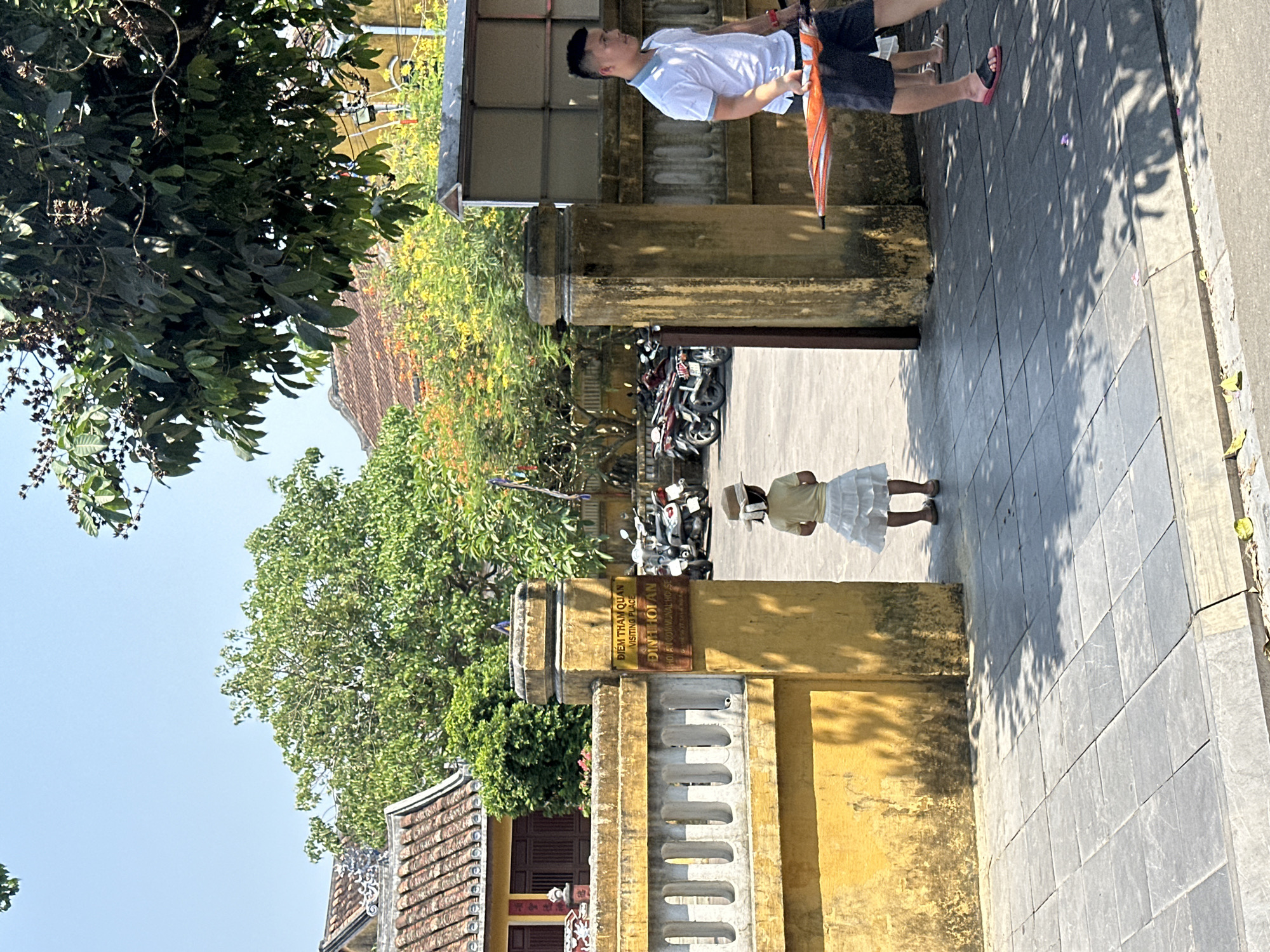 A vertical, medium shot in daylight captures a street scene in Hội An Ward, Vietnam. The street is paved with gray tiles, and on the right, the edge of a sidewalk is visible. A man in a white shirt and dark shorts, holding a striped umbrella, walks along the sidewalk. To the left, an entrance to a building is framed by weathered yellow walls. A small sign on the wall reads "ĐIỂM THAM QUAN ĐÌNH HỘI AN", denoting a visiting place. A young girl in a light-colored dress and hat stands in the entrance. Behind her, a row of motorbikes is parked. Above, lush green trees and foliage overhang the scene, with a glimpse of a traditional tiled roof of a building in the background. The sky is a clear blue.
