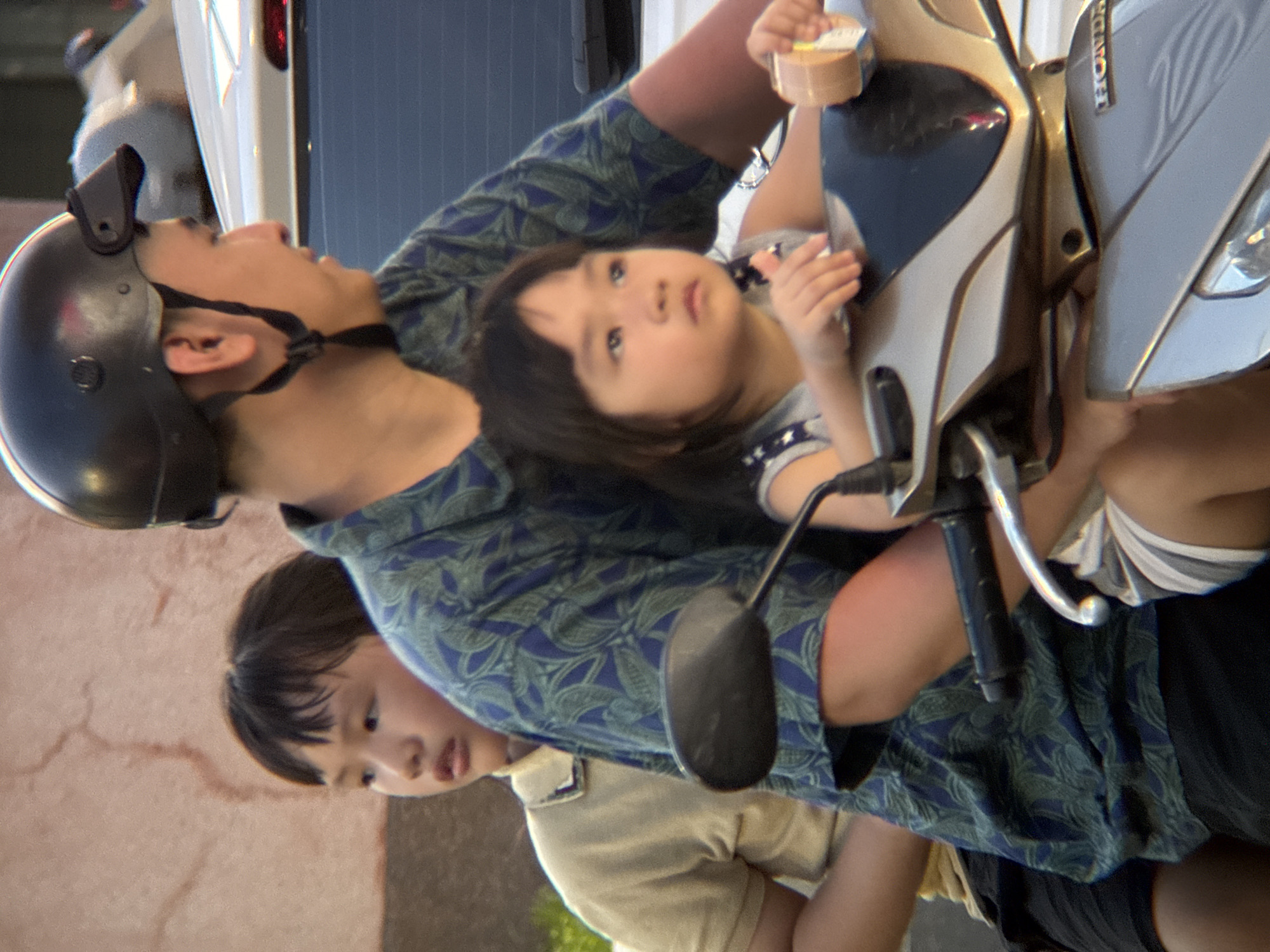 A family of three rides on a motorcycle in Nha Trang, Vietnam. The father, wearing a black helmet and a blue and green patterned shirt, is driving. A young boy, wearing a tan polo shirt and black shorts, sits behind him. A young girl, wearing a gray and white striped shirt, sits in front of the father. The motorcycle is silver and gray, with a visible "HONDA" logo on the front. The background appears to be a street scene, with a building wall and another vehicle visible. The lighting suggests it is daytime, possibly late afternoon, with warm, soft light.
