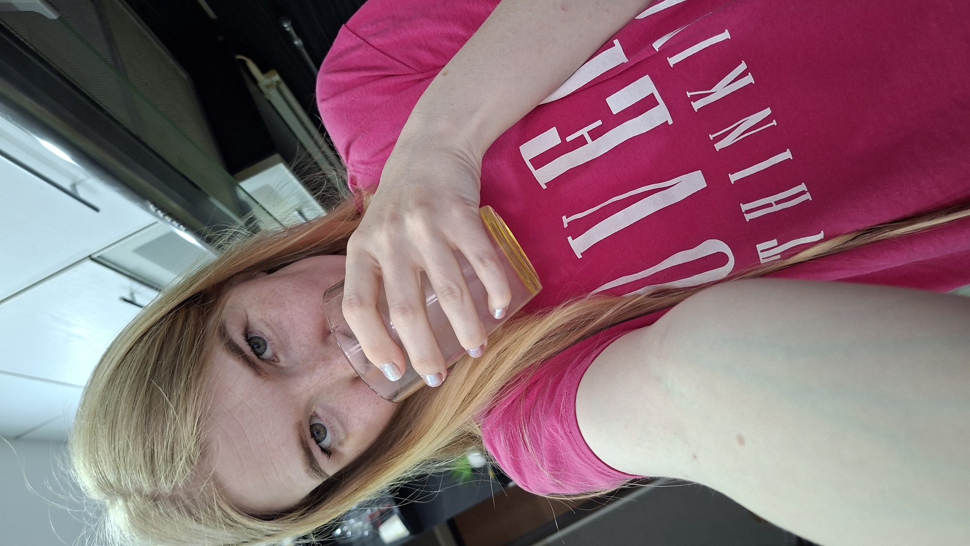 A blonde woman with fair skin is holding a glass up to her face. She has freckles, green eyes, and wears glasses. She is wearing a bright pink t-shirt with white lettering. The scene appears to be indoors, possibly a kitchen, with overhead cabinets visible in the background. The exact location within Chaikovsky, Russia, cannot be determined from the visual data provided.