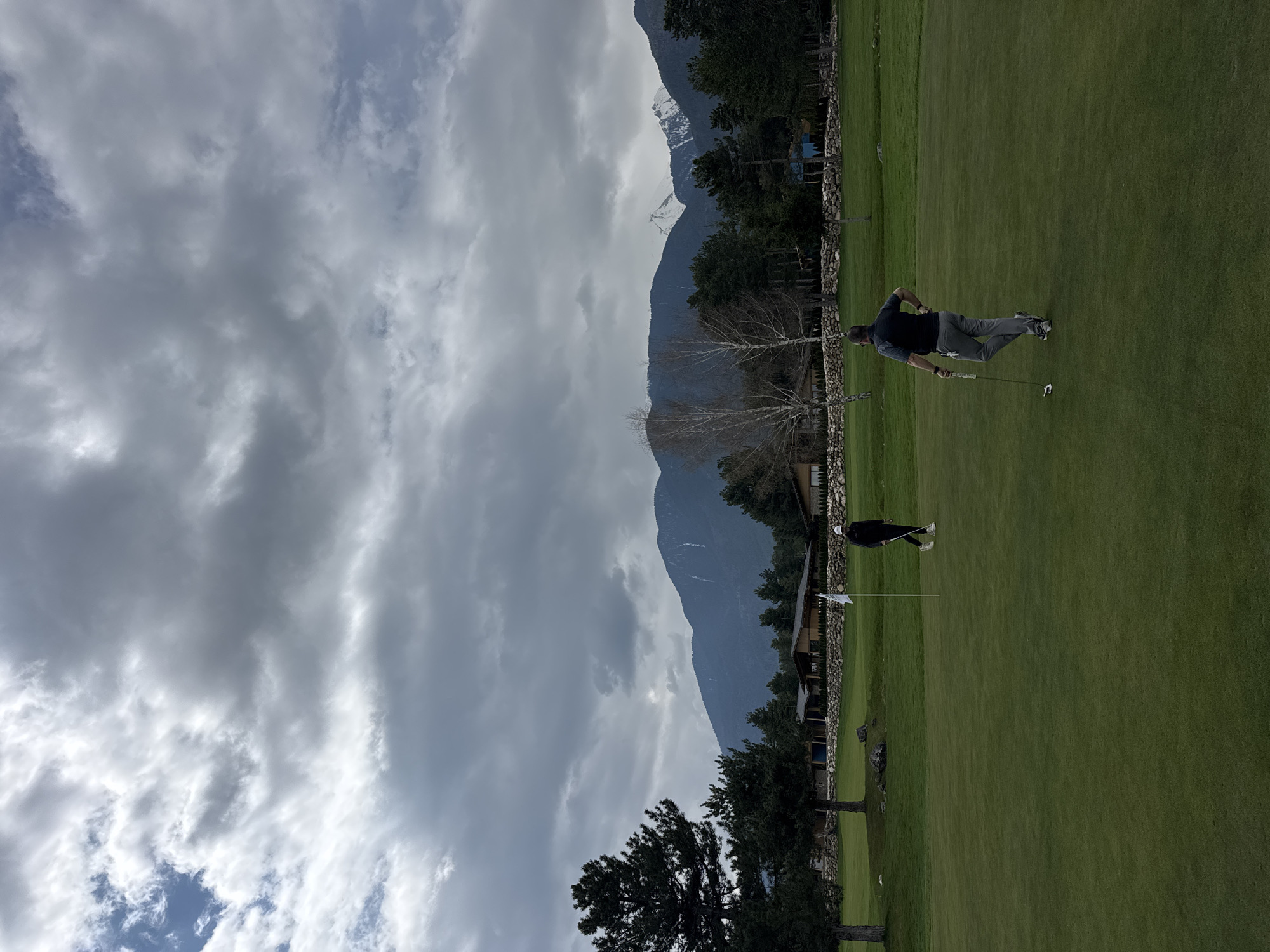This media file captures a golf course scene set against a dramatic mountainous backdrop. The primary subjects are two men engaged in golfing activities on a well-maintained green. One man, dressed in a dark shirt and grey pants, is positioned towards the center-right, holding a golf putter, appearing to be in the process of putting or having just completed a stroke. Further back, another person in dark attire and a white hat stands near a white flagstick, which marks the hole on the green.
The setting is characterized by lush green fairways and a distinctive stone wall that runs across the mid-ground. Behind this wall, a mix of trees, including some deciduous trees with bare branches and evergreen conifers, are visible. In the background, majestic mountains rise prominently, with some peaks displaying patches of snow, suggesting a high altitude or cooler season. The mountains are rugged and appear to have forested slopes.
The sky is largely overcast with significant grey cloud cover, although glimpses of lighter clouds and patches of blue sky can be seen, indicating daytime. The light is diffused due to the clouds. A building or structure, possibly part of a golf club or accommodation, is partially visible behind the trees and stone wall on the right side of the image. The scene conveys a peaceful atmosphere, typical of a golf course in a scenic, mountainous region like Razlog, Bulgaria. There is no visible text in the image.