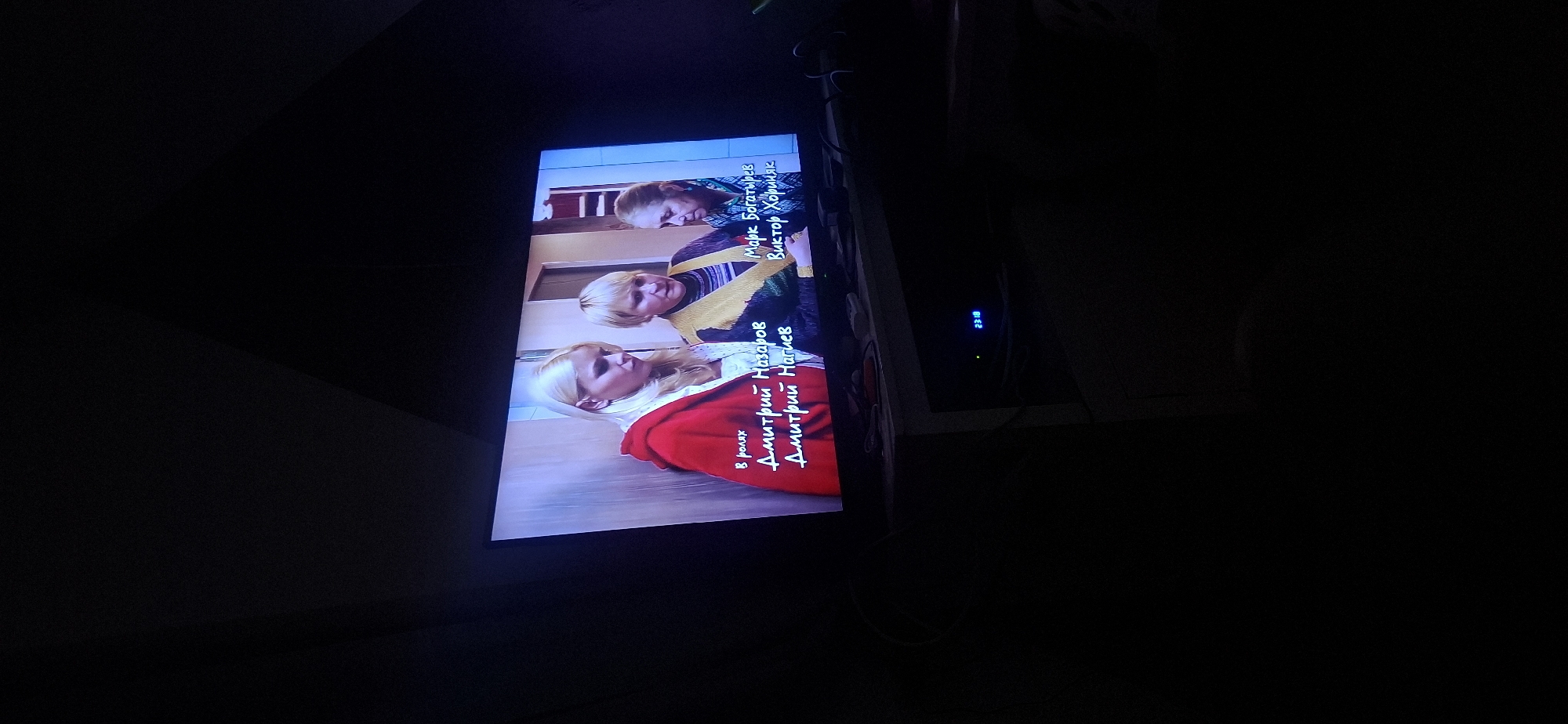 The image shows a television screen displaying a scene from a film or television show, with text overlays indicating actors' names. The screen is positioned on a dark furniture unit. The lighting in the room is dim, with the television screen being the primary light source. The overall scene suggests an indoor setting at night, possibly a living room, where someone is watching television. There is no indication of the city of Yerocham, Israel, in the image.