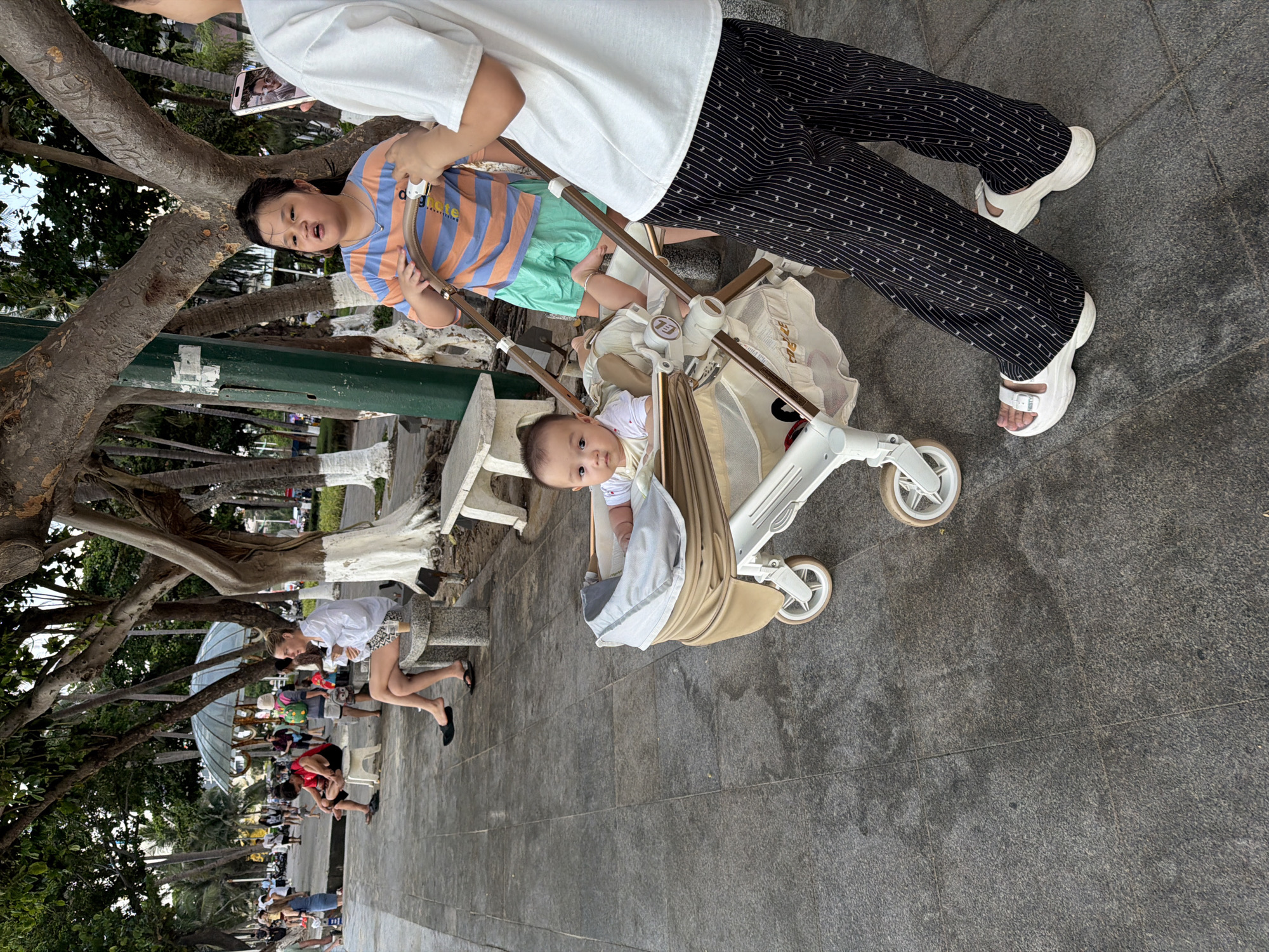 A group of people are gathered on a paved walkway lined with trees and palm trees. In the foreground, a person wearing black and white striped pants and white sandals pushes a light-colored stroller. Inside the stroller is a baby looking at the camera. Next to the stroller, a child in an orange and blue striped shirt and green shorts looks toward the phone being held by a person whose upper body is visible, wearing a white t-shirt. In the background, other people are visible walking and sitting. One person in a white shirt and patterned shorts is bending down. The scene appears to be in a park or along a promenade, possibly in Nha Trang Ward, Vietnam, given the lush vegetation and architecture. The lighting suggests it is daytime, and the atmosphere seems relaxed, with people enjoying an outdoor setting. No visible text is present in the image.