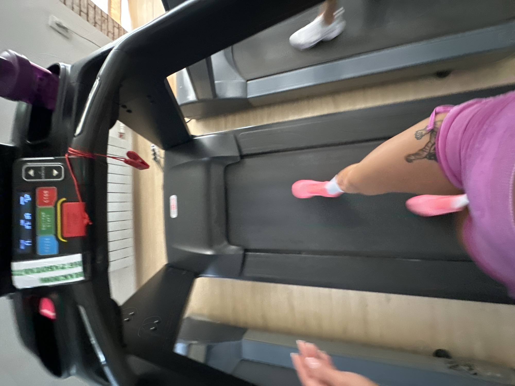 An individual, identified by their pink athletic shoes and a pink garment, is exercising on a treadmill. The individual's left leg, adorned with a tattoo of a tiger, is visible. The treadmill is positioned indoors, likely in a gym or fitness center, with other treadmills visible in the background. The environment appears to be clean and well-lit, with wooden flooring. The image does not provide any specific information to determine its location within the city of Yuzhnouralsk, Russia.