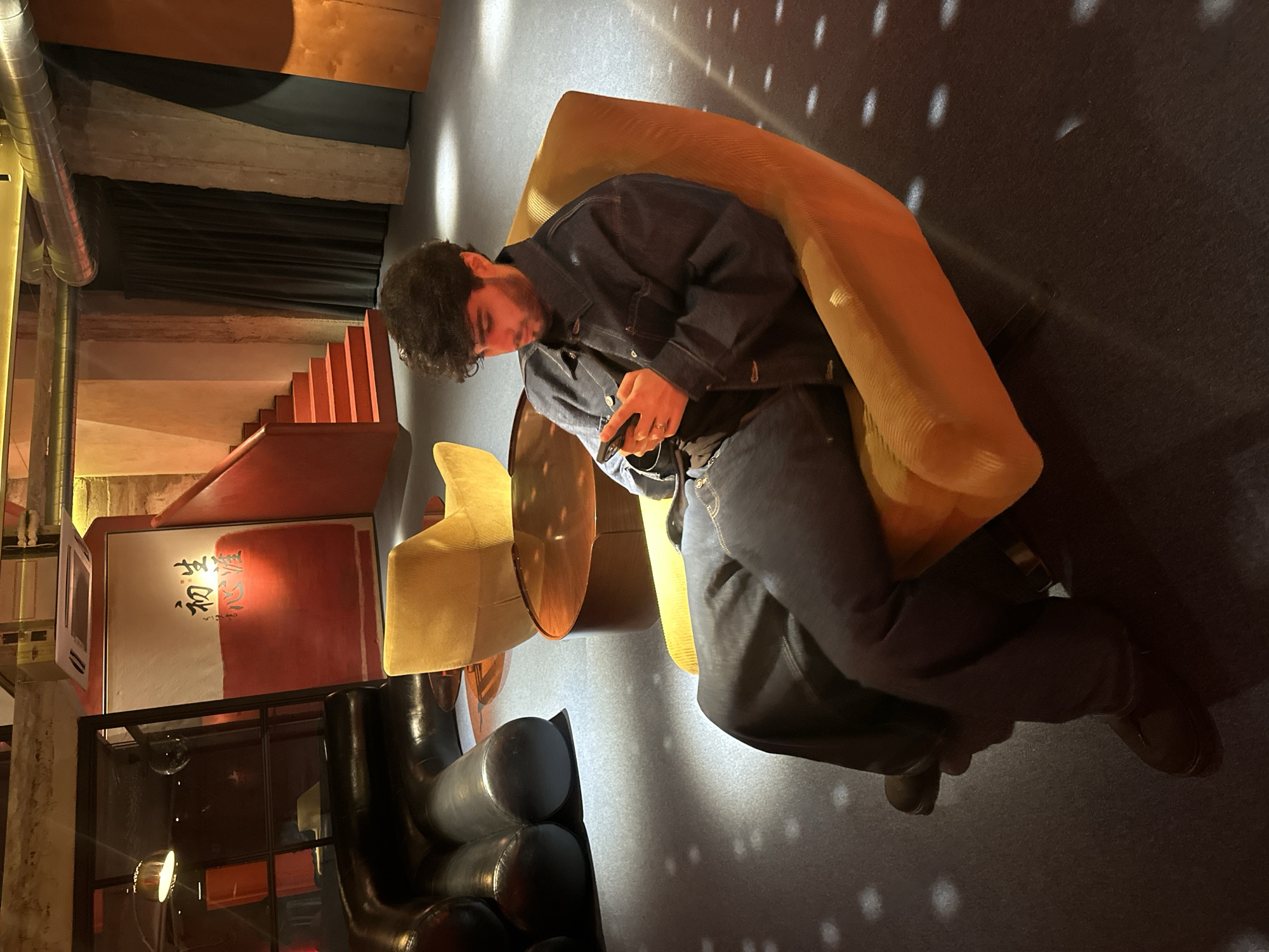 A medium, angled shot shows a man seated in a yellow chair indoors, possibly in Yerevan, Armenia. The man has dark, curly hair and a beard, and he is wearing a denim jacket and jeans. He is looking down at a phone in his hands. The chair is cushioned and appears to be part of a modern interior design. The floor is a dark gray carpet.
In the background, there is a staircase with red steps, a black curtain, and a piece of art with red and white paint and black characters. There are also some decorative elements and a metal pipe. The lighting is dim, with the primary light source coming from overhead. The overall scene gives an impression of a stylish, contemporary space, possibly a restaurant or lounge.