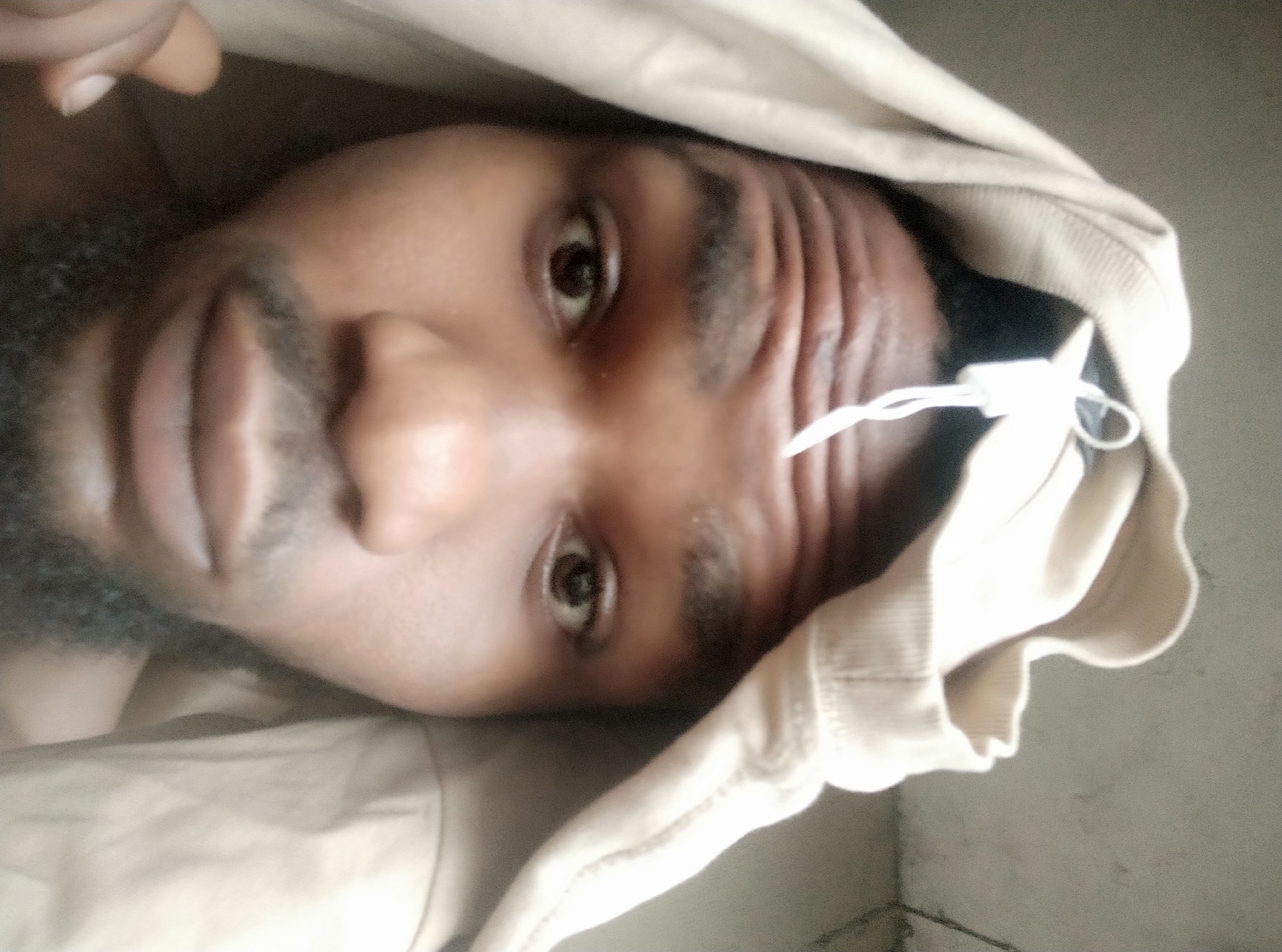 A medium shot of a bearded man in a tan hooded sweater is lying down. His head is covered by the hood and he looks directly at the camera. The location is Unknown, Unknown.