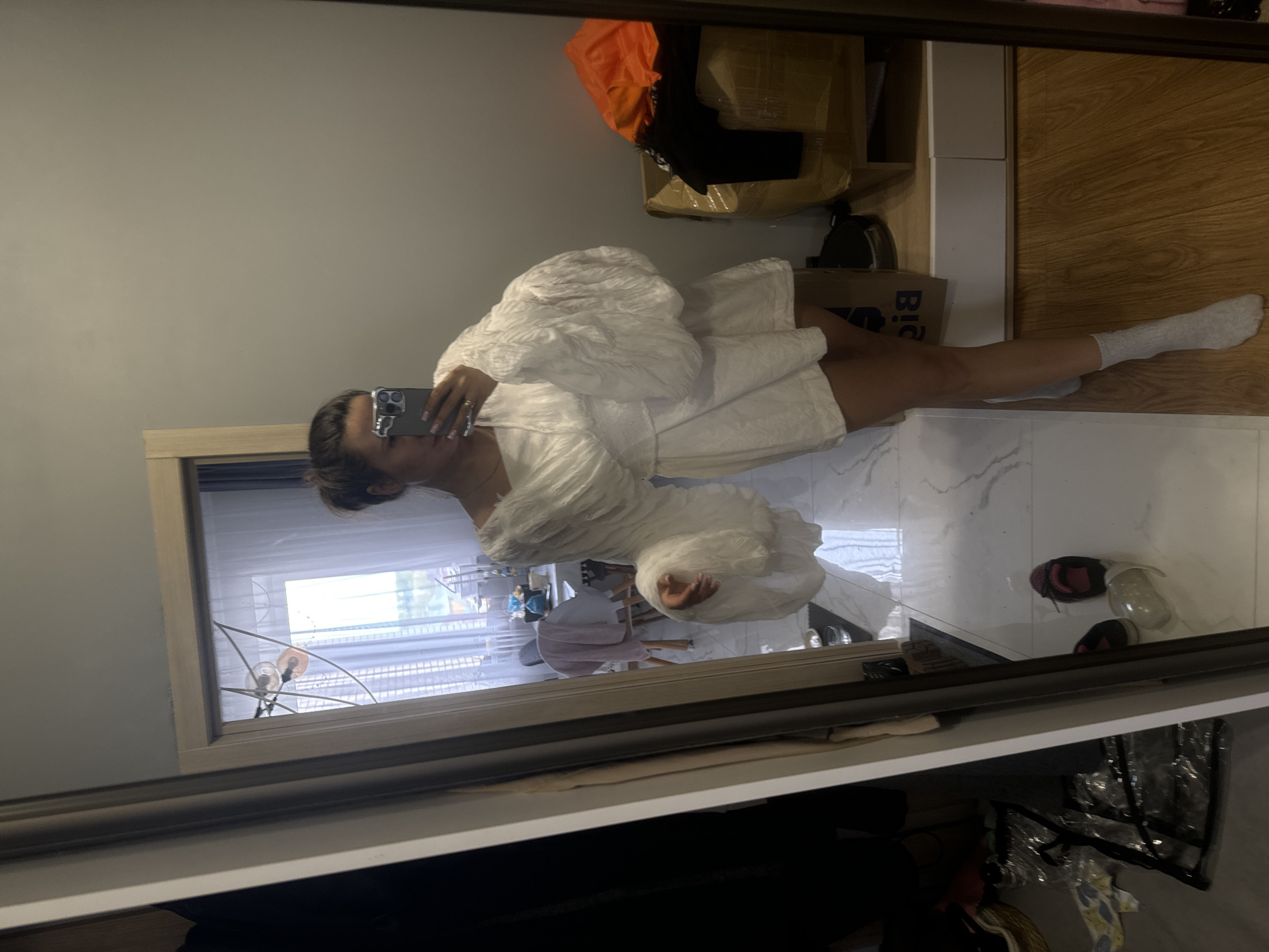 A person is reflected in a mirror, holding a cell phone to take a selfie. The person is wearing a textured white outfit with puffed sleeves and white socks. The room features a window with daylight coming through, a wooden floor and shelf, and a marble-patterned floor. There are various items visible, including packages, shoes, and clothing, scattered in the periphery of the image. There is insufficient information to determine if this image was taken in the city of Odintsovo, Russia.