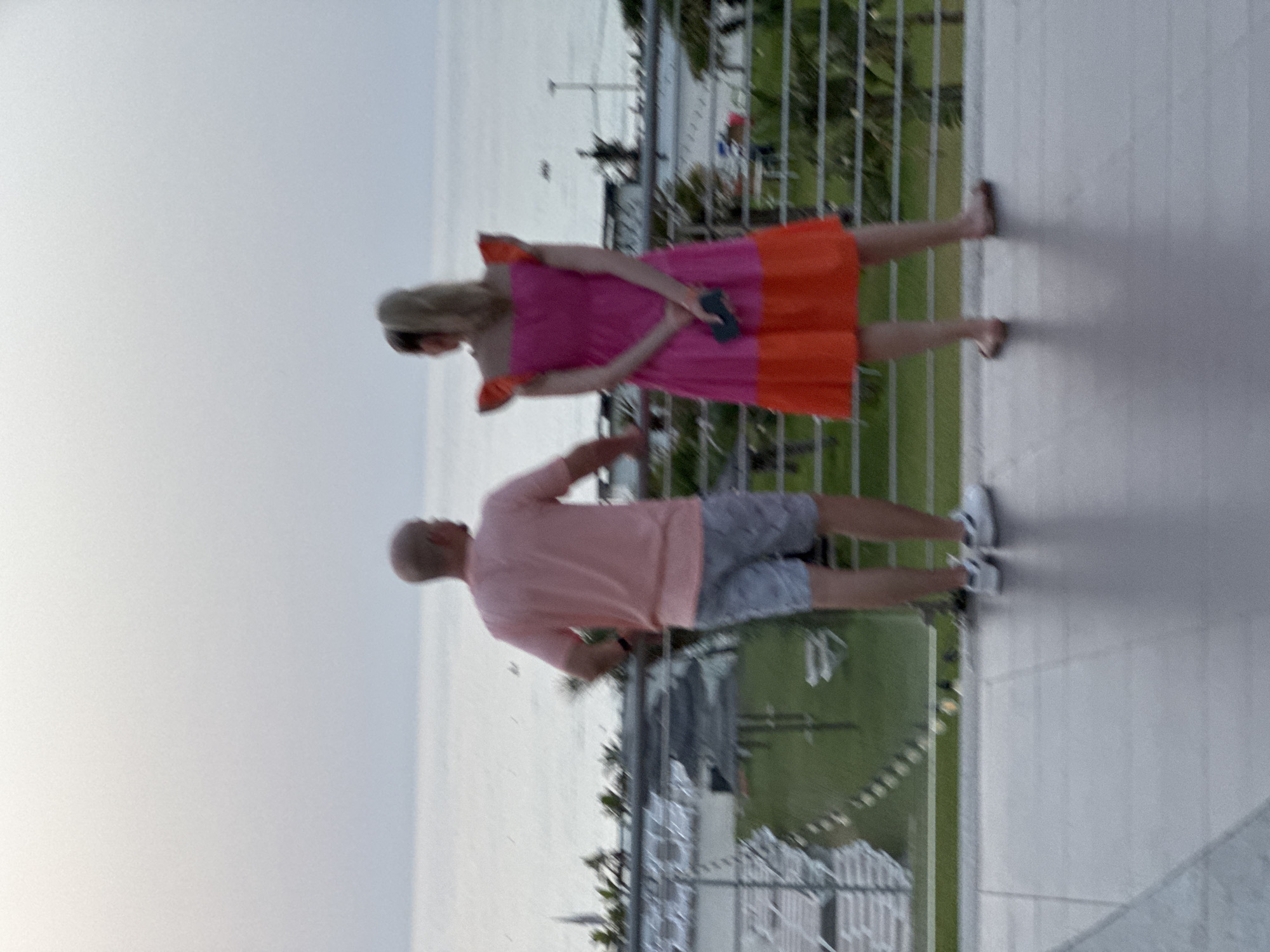 The image shows a man and a woman standing on a balcony overlooking the sea. The man is wearing a pink t-shirt and gray patterned shorts. The woman is wearing a pink and orange two-tone dress with ruffled sleeves. She is holding a dark-colored phone in her right hand. In the background, the sea is visible, with a coastline and some buildings in the distance. The scene appears to be set in Capaci, Italy, given the context of the prompt. The overall atmosphere is relaxed, with the subjects appearing to be enjoying the view.