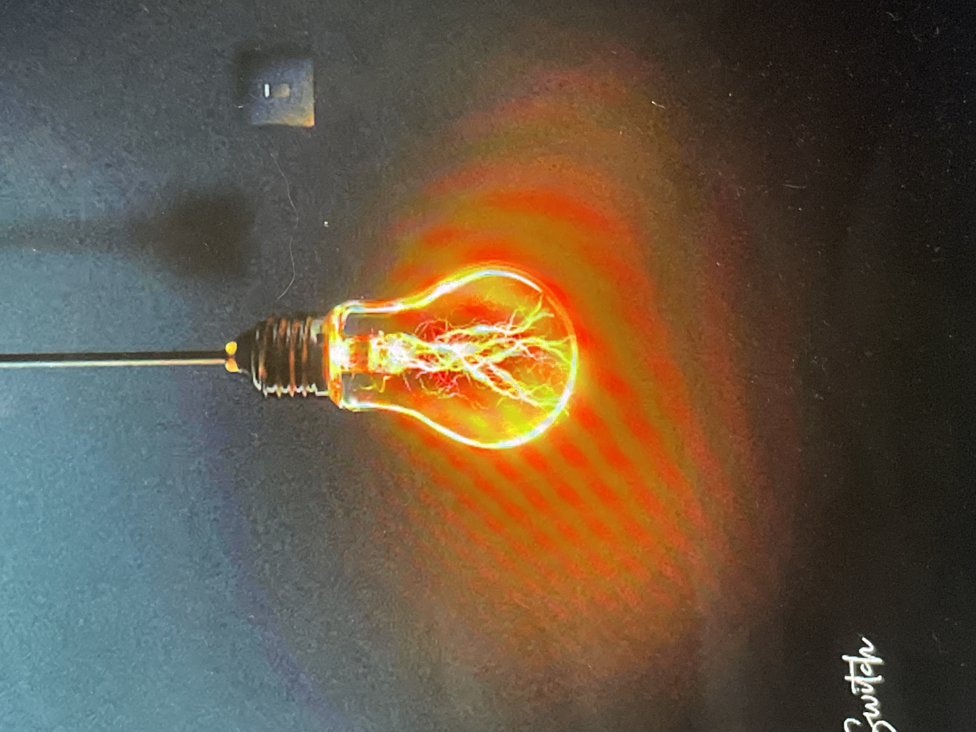 This media file features a glowing, plasma-like light bulb set against a dark background, likely captured indoors in Sofia, Bulgaria. The central subject is a decorative light bulb, resembling a traditional incandescent bulb, filled with bright, branching, yellow-white electrical discharges that create a plasma effect. These discharges emit a vibrant orange and yellow light, which radiates outwards in a concentric pattern around the bulb, illuminating its immediate vicinity.
The bulb is positioned horizontally and appears to be connected to or held by a slender, dark stick or rod extending from the left side of the frame. The background is a uniform dark surface, possibly a wall or screen, with visible dust particles and fine fibers scattered across it. In the upper-left corner, a small, dark rectangular object with two light-colored horizontal slots is visible, resembling a wall outlet or switch plate. Below this, a faint, dark, smudged area is discernible on the background.
In the bottom-right corner of the image, the word "Switch" is visible, rotated ninety degrees clockwise, rendered in a stylized, light-colored font. The overall impression is of a scientific or artistic display, with the intense luminescence of the bulb being the primary focal point, captured in a dimly lit or dark environment to enhance its glow.