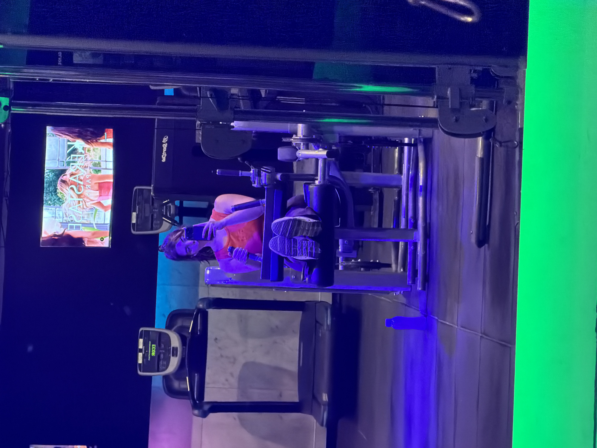 This image, taken in Buenos Aires, Argentina, captures an indoor gym or fitness center environment, characterized by dim lighting and vibrant blue and neon green accent lights.
The primary subject is a young woman, seen from a side profile, seated on a piece of dark-colored exercise equipment, possibly a leg press machine. She is wearing an orange top and sneakers with visible textured soles. Her activity involves holding and looking at her smartphone, suggesting she is either taking a break from her workout or waiting to begin.
To her left, a large screen displays what appears to be a scene from a television show or movie, featuring two women—one with dark hair and one with blonde hair—in an outdoor setting with a white fence. The word "PARIS" is partially visible on this screen. Another screen, attached to a piece of exercise equipment in the foreground, displays the text "E006". A dark blue water bottle rests on the floor beside the woman's machine.
The overall atmosphere is modern and somewhat moody due to the artificial lighting. Other mechanical components and structures of gym equipment are visible throughout the scene. Visible text includes "PARIS" on the main display, "E006" on the foreground machine's screen, and "ORIDE" (upside down) on a panel in the upper left, potentially part of a brand name. A stylized logo resembling "PROXIMA" can also be seen on the side of the equipment the woman is using.