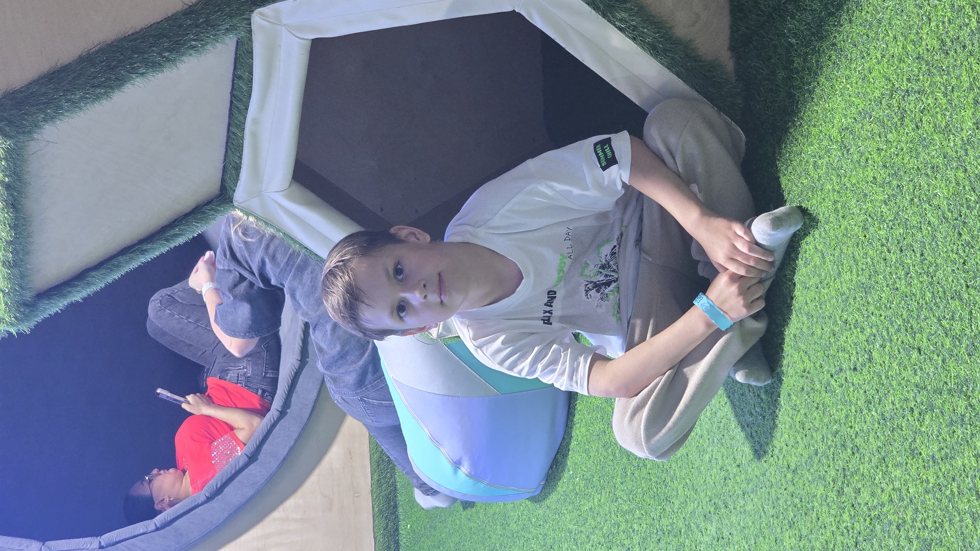 The image depicts an indoor recreational area. The floor is covered with bright green artificial turf.
In the foreground, a male child, estimated to be between 8 and 12 years old, sits cross-legged on the turf, facing the camera with a neutral expression. He wears a white short-sleeved T-shirt with the text "FLAX AND ENJOY ALL DAY" and a palm tree graphic, light brown trousers, and grey socks. A light blue wristband is visible on his left wrist.
In the mid-ground, to the left, an adult female is reclining within a grey, curved, padded seating structure. She is wearing a red short-sleeved top with embellishments, dark jeans, and glasses, and is holding and looking at a smartphone or similar electronic device.
Also in the mid-ground, partially obscured behind the child and beneath a white, tent-like structure trimmed with artificial turf, the lower torso and legs of a second adult are visible. This individual wears a dark long-sleeved top and dark jeans, with a white wristband on the left wrist, and appears to be in a reclining position on the turf.
Behind the child, a multi-colored (light blue, white, and light green) segmented beanbag or pillow rests on the artificial turf. The tent-like structure to the child's right has a cream or white frame with a green artificial turf trim along its top edge. A light wooden panel wall is visible behind the grey seating structure.
No specific visual markers within the image identify the location as Izhevsk, Russia.