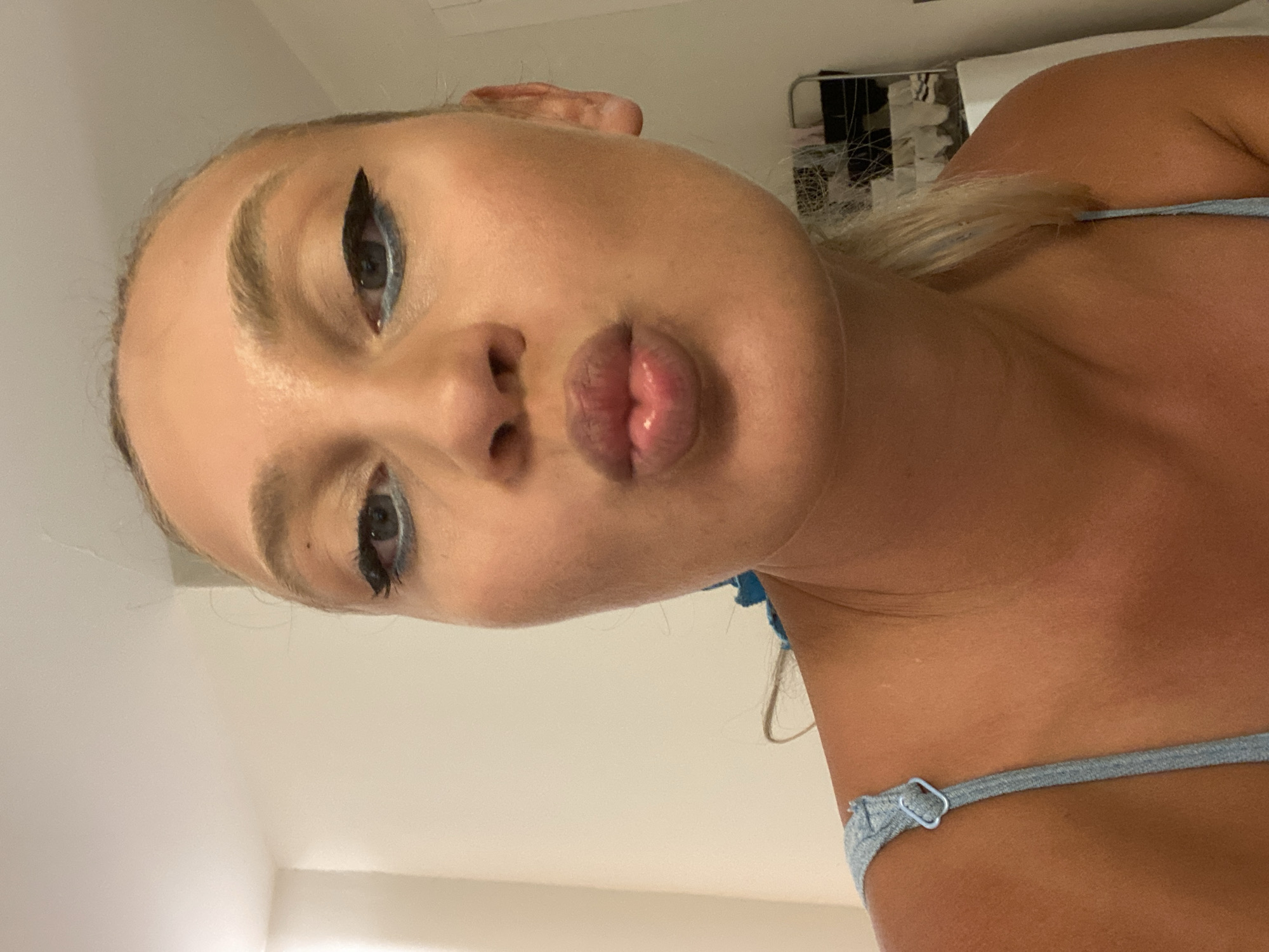 The image shows a close-up of a young woman's face, with her lips pursed in a kissing gesture. She has blonde hair and is wearing a light blue top with a strap visible over her shoulder. Her makeup includes winged eyeliner and blue eyeshadow. The background is a plain, light-colored wall. The image is a selfie, taken from a low angle. There is no visible indication of the city of Aksu, Turkey, or any specific urban environment.