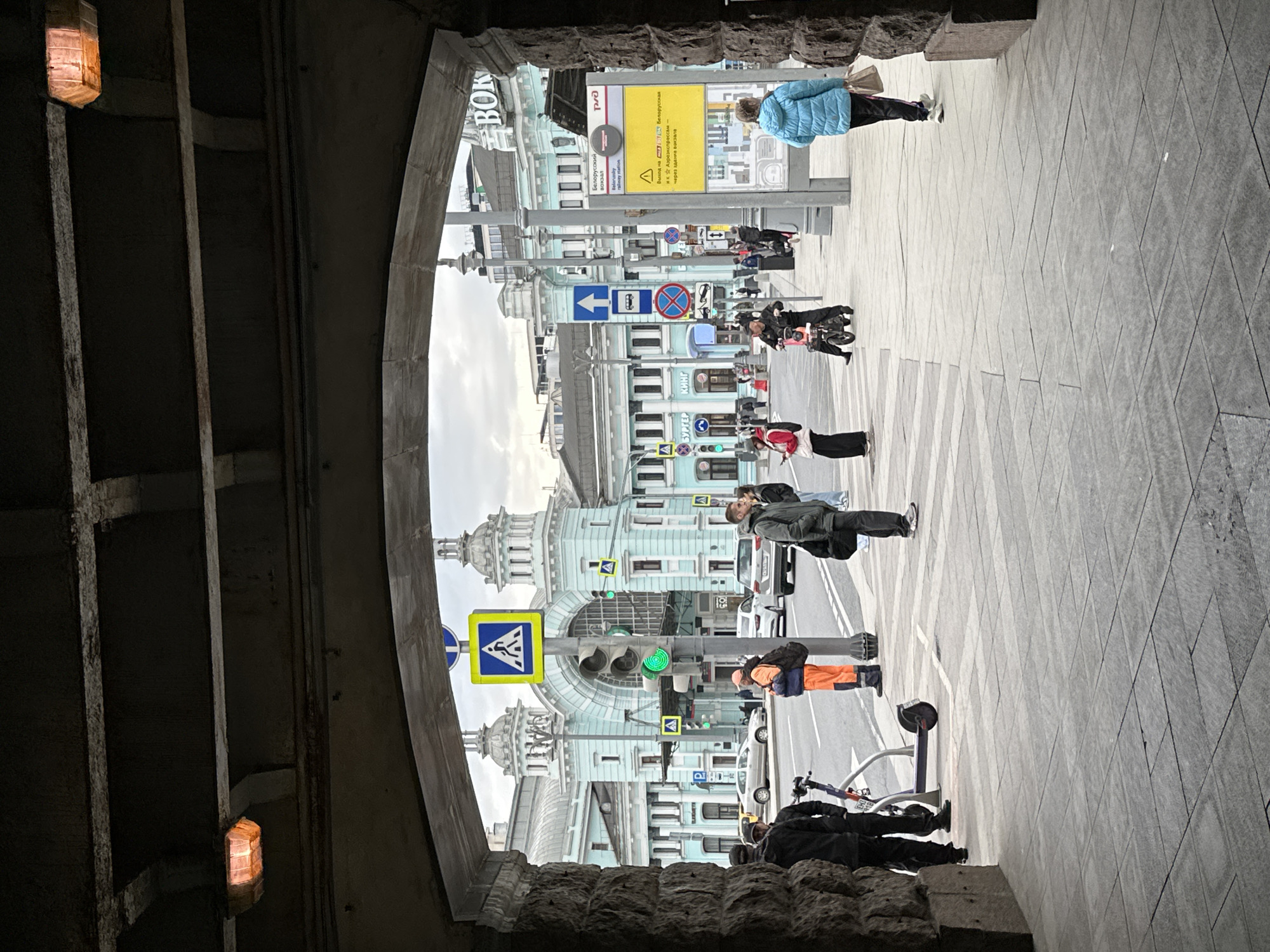The image shows a street scene in Moscow, Russia, viewed through an arched opening, possibly a pedestrian underpass. The architectural style of the buildings suggests a historical area. People are walking on the sidewalk. Road signs and traffic lights are visible, including a pedestrian crossing sign and a traffic light showing a green light. A sign indicates "Belorussky Station" and "Aeroexpress". A pedestrian wearing an orange work outfit is visible. Bicycles and scooters are along the sidewalk. The overall light suggests an overcast day.