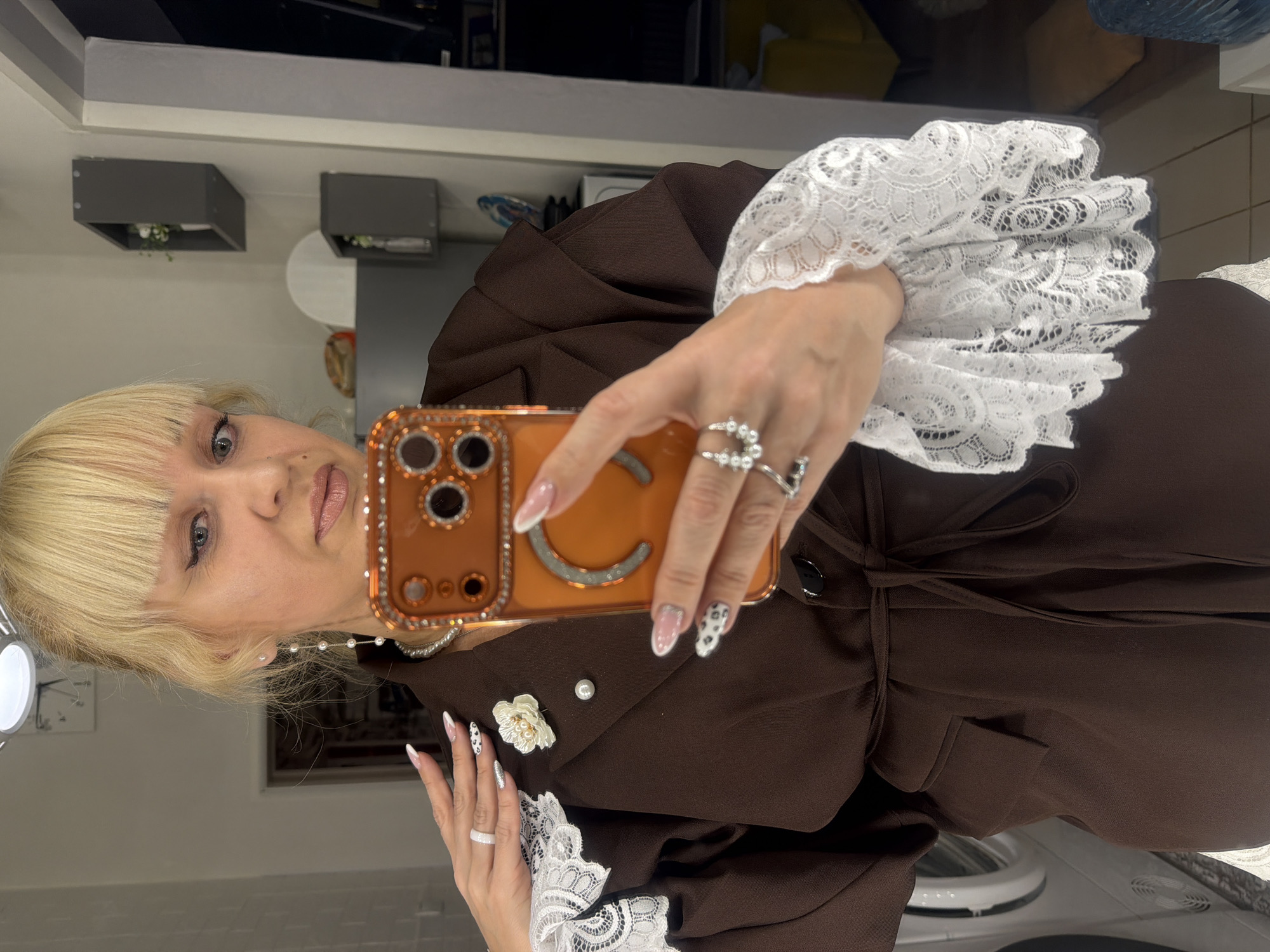 A person with blond hair and bangs is taking a selfie with an orange phone case. The person is wearing a brown jacket with a tie belt and a white lace-trimmed cuff. The person has rings on their fingers and pearl earrings. The background appears to be a room with cabinets and a washing machine. The location of the image cannot be verified.