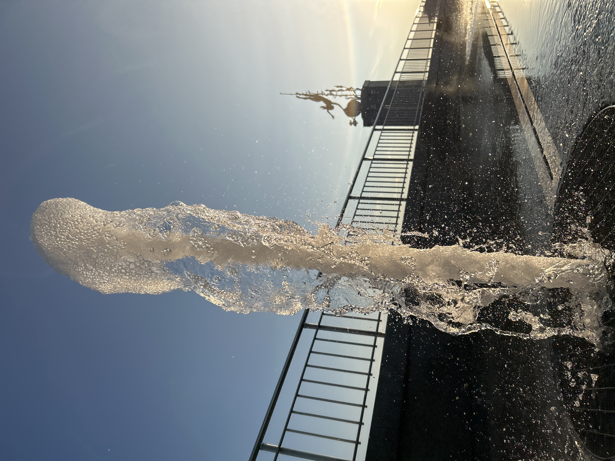 This image captures a close-up view of a water jet erupting from a fountain, set against a bright, clear sky and a dark, reflective surface. The water forms a dynamic, frothy stream that dominates the left side of the frame, with numerous droplets catching the sunlight. In the background, a tall, dark structure with horizontal lines, possibly a building or a modern sculpture, is visible. A metallic railing runs along the edge of this structure, leading upwards. At the top of the structure, a silhouette of a sculpture featuring figures in mid-leap is discernible against the bright sky, which appears to be bathed in the warm glow of the sun, suggesting it is either sunrise or sunset. The overall scene evokes a sense of motion and the refreshing presence of water. The location is Tolyatti, Russia.