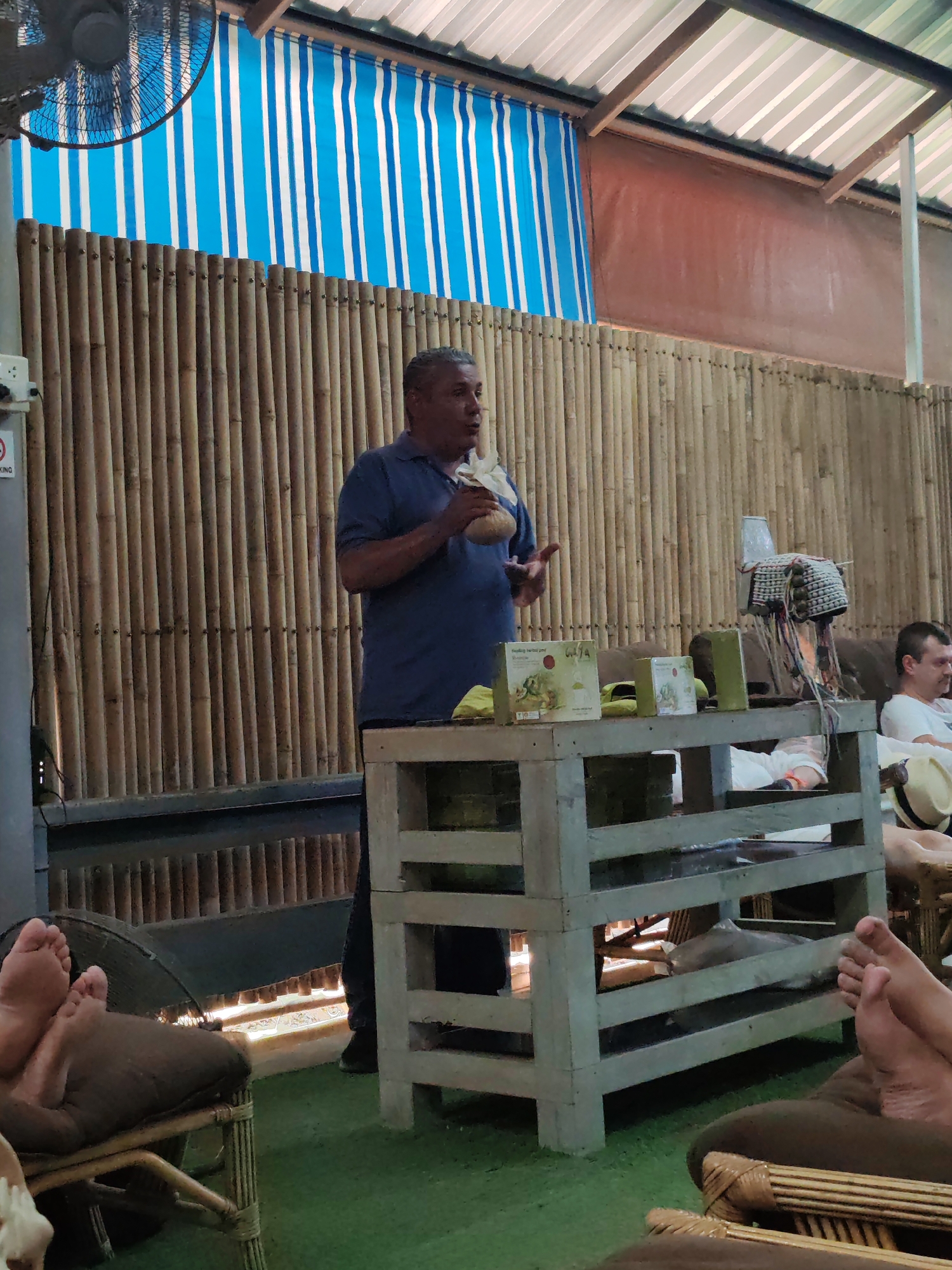 This image captures a relaxed, semi-open-air setting, likely a wellness center or shop in Ban Khlong Khian, Thailand. The primary subject is a middle-aged man with short, greying hair, wearing a blue polo shirt. He stands in the center-left, facing right, holding a light-colored, natural material item (possibly a small woven basket or coconut shell) in his left hand and gesturing with his right, appearing to explain or present something to an unseen audience.
The background features a tall wall constructed from vertical bamboo poles, lending a rustic, natural aesthetic. Above the bamboo wall, a blue and white striped awning provides shade, and further up, a corrugated metal roof is visible, supported by wooden beams. An industrial-style fan hangs from the ceiling in the upper left.
In the immediate foreground, several pairs of bare feet are visible, resting on cushions or low chairs, indicating patrons are comfortably seated and relaxed, possibly receiving a foot massage or enjoying a spa treatment. To the right, another person is partially visible, sitting in a chair.
A rustic, light-colored wooden table or shelving unit stands in front of the speaker. On this table are several green product boxes; one is clearly labeled "Healing Herbal pad," and text like "CASA" or "GASA" is partially visible on another. A decorative, woven item with dangling threads and a yellow cloth also rest on the table. A partial "NO SMOKING" symbol is visible on a power outlet on the left wall. The floor appears to be covered with green artificial turf. The overall atmosphere suggests a casual, informative session in a tropical, relaxed environment.