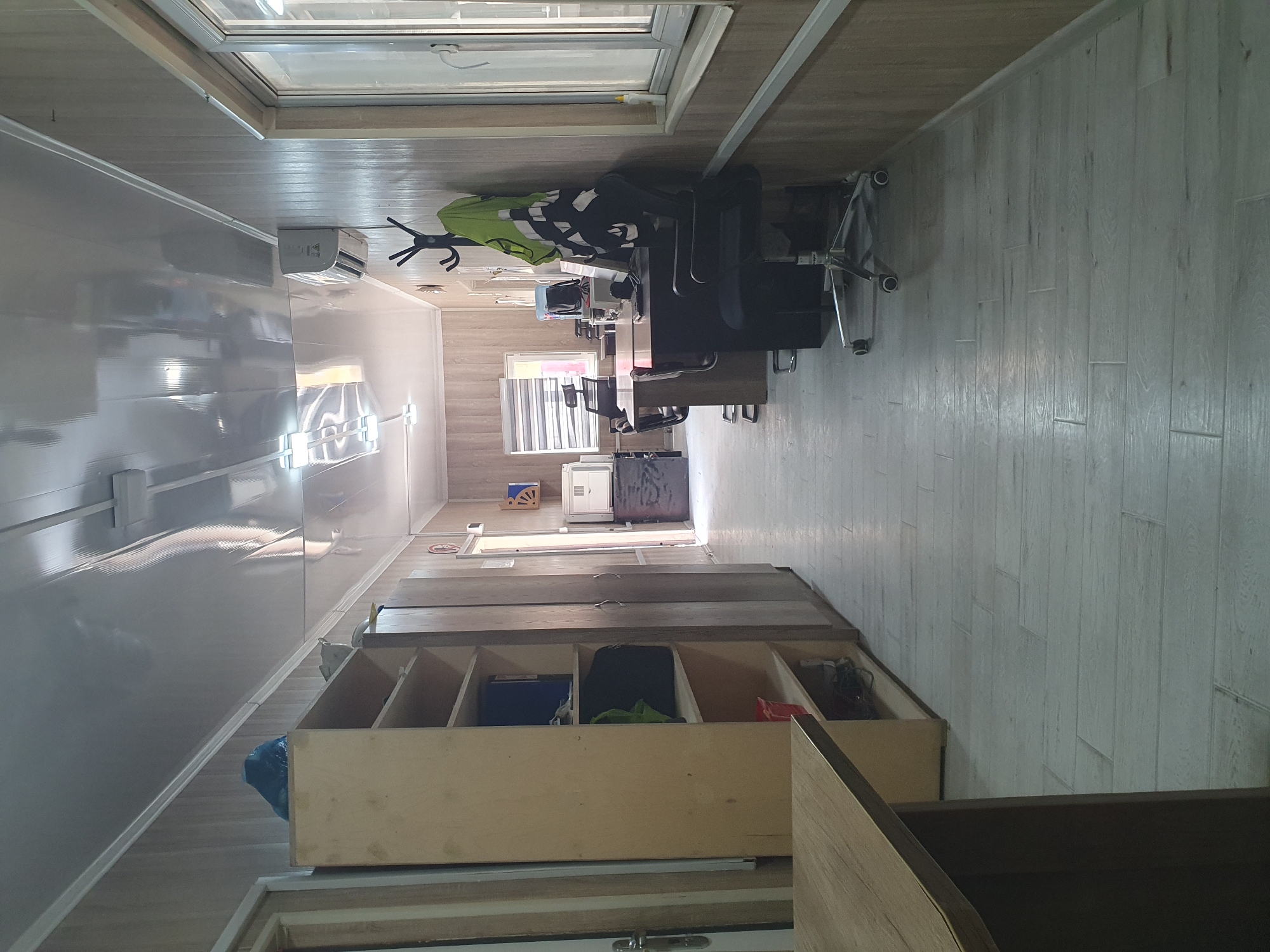 The image depicts a long, narrow office space with light-colored wooden paneling on the walls and a reflective, white ceiling. The floor is made of light-colored wooden planks laid in a staggered pattern. The space appears to be a portable office or a modular building, as suggested by the construction and the overall aesthetic.
On the left side of the image, there is a tall, light-colored wooden shelf unit filled with various items, including bags and containers. A dark blue garbage bag is placed on top of the shelf. Further down the corridor, more wooden cabinets are visible.
The right side of the image features a window with blinds, letting in natural light. Adjacent to the window are several office desks with computers and office chairs. A black office chair with wheels is prominently placed in the foreground. A high-visibility jacket in green and black is hanging on a coat rack.
In the background, there are more desks and office equipment, including what appears to be a printer and a small refrigerator. An air conditioning unit is mounted on the wall above one of the desks. The overall impression is one of a functional, if somewhat utilitarian, workspace. There are no people visible in the image, and no specific details are discernible that would allow for pinpointing the exact location within Tashkent, Uzbekistan. However, the style of construction and the presence of office furnishings are consistent with commercial or industrial settings found in the city.