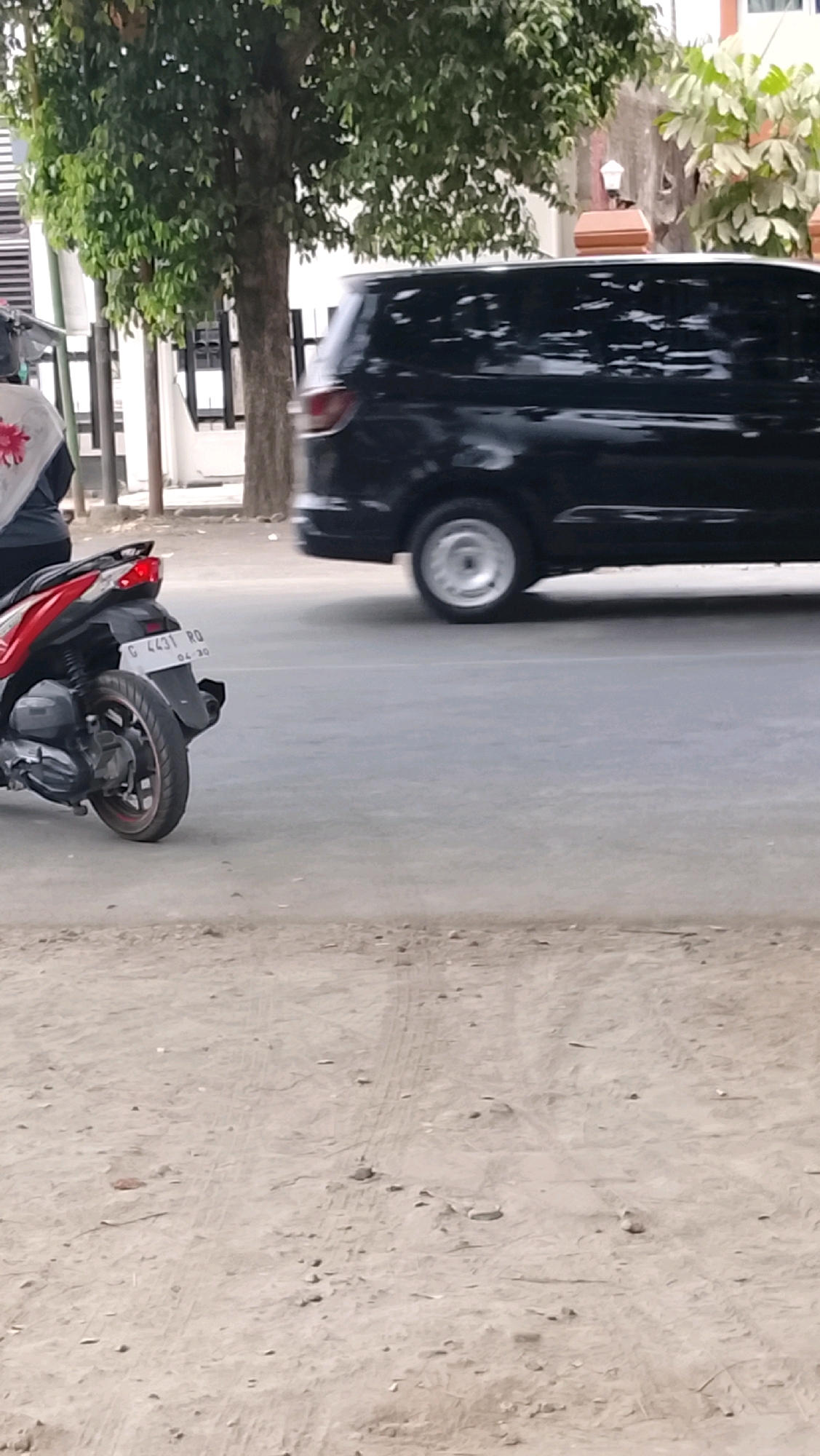 An outdoor, eye-level, medium shot shows a black minivan and a red and black motorcycle on a paved road. The minivan is to the right, in motion, with its front end cut off. The motorcycle is to the left, stationary, and angled towards the right. The rear tire of the motorcycle has a red rim and is partially obscured by the vehicle’s body. The motorcycle’s license plate reads “G 4431 RQ” with smaller numbers below it. In the background, on the left, a person is partially visible, wearing a white garment with a red floral design. A mature tree with green leaves stands behind the person, and further back, a white building with a black fence is visible. To the right of the minivan, a portion of a building with orange and brown elements is seen. The foreground consists of a dirt or gravel surface. The scene appears to be a street in Slawi, Indonesia.