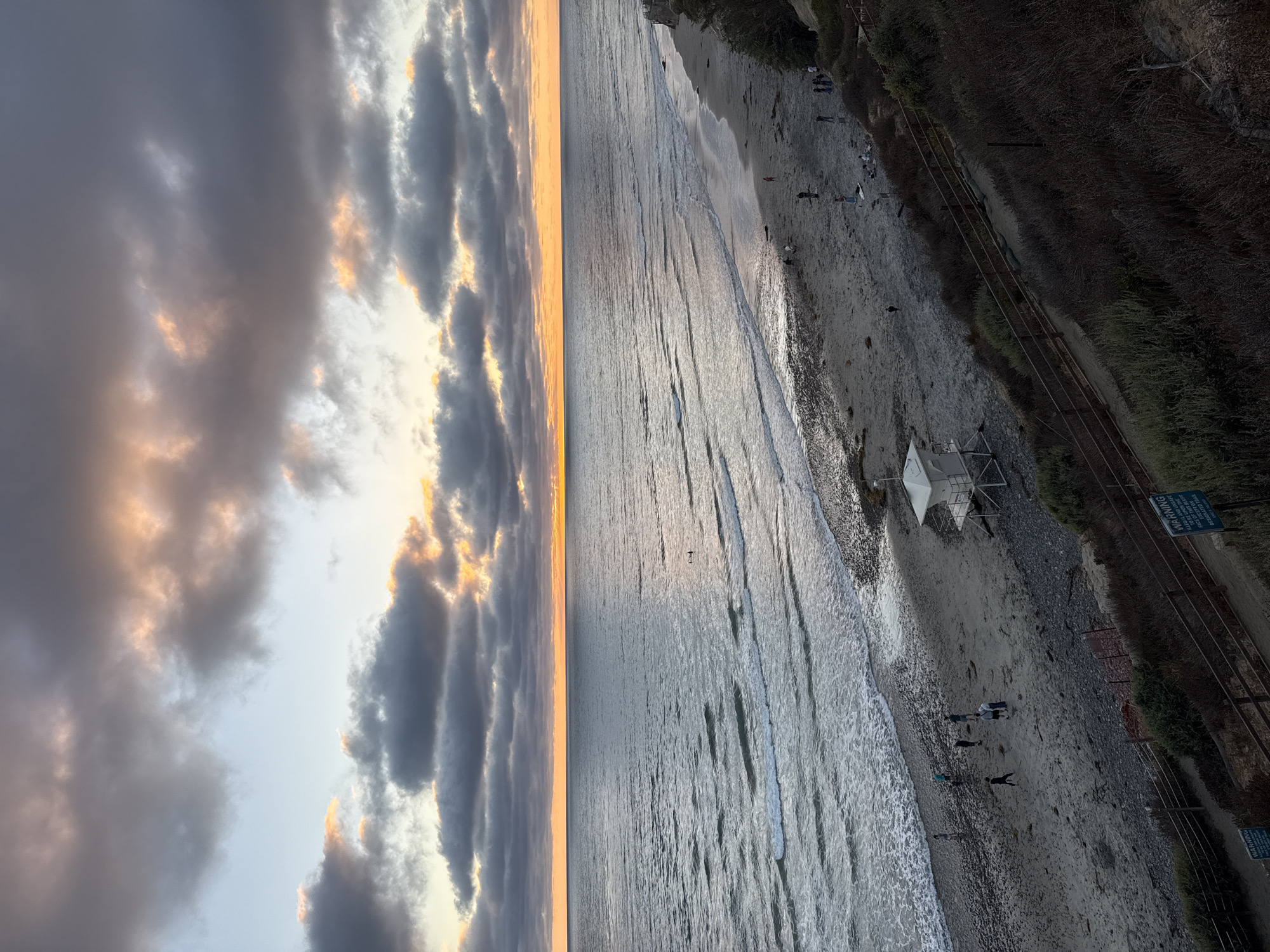 A wide-angle, elevated shot captures a beach scene at dusk in Encinitas, California. The sky is filled with dramatic, overcast clouds illuminated by the setting sun, which casts a golden glow along the horizon. The ocean stretches out, with gentle waves rolling onto the sandy and pebbly shore. Several individuals are present on the beach, some walking, others gathered in small groups. A white lifeguard tower stands on the sand near the water's edge. A winding pathway or stairs, bordered by vegetation and a wooden fence, leads down from an elevated vantage point, where the photograph was taken. The overall mood is serene and picturesque.