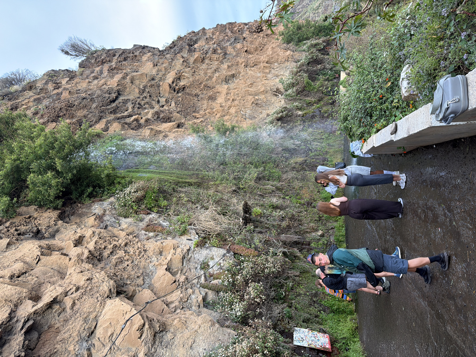 The image depicts a group of people standing in front of a waterfall cascading down a rocky cliff face, likely located in the city of Ponta do Sol, Portugal. The cliff face is primarily composed of light brown and dark brown rocks, with some patches of green vegetation clinging to the surface. The waterfall flows from the top of the cliff, creating a misty spray as it descends.
In the foreground, four individuals are present. Two are a couple with the man in a green jacket and the woman with long brown hair. In front of them are another man, who seems to be taking a picture with his phone, and a woman wearing a grey shirt. A concrete walkway or ledge is visible, with a grey backpack or bag resting on it near the edge. Green bushes and small white flowers grow along the edge of the walkway. The ground appears wet.
To the bottom left, there is a mosaic table, adding a touch of art to the natural setting.