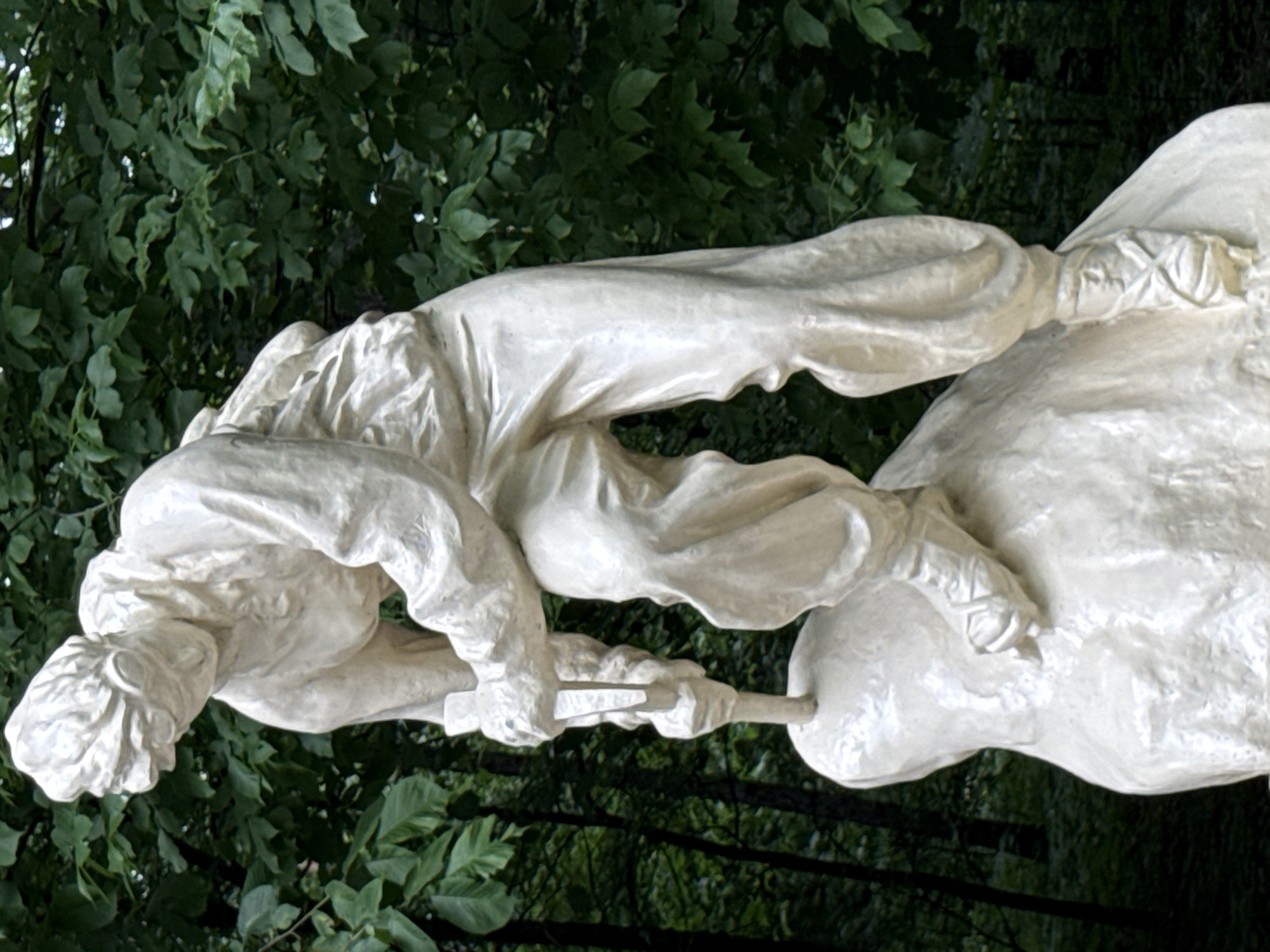 This is a close-up, rotated, outdoor photograph of a white, classical-style sculpture of a man against a backdrop of dark green foliage. The sculpture appears to be made of stone or a similar material and depicts a person in dynamic motion. The figure is lean and appears to be kneeling or bending over a large, rounded object. Their left arm is extended, and they are holding a tool or implement that is in contact with the object. The figure wears clothing that drapes in flowing folds, suggesting a historical or stylized representation. The fabric details are highly rendered, with deep crevices and smooth surfaces. The head of the figure is visible, with sculpted hair and facial features, although the resolution makes fine details difficult to discern. The overall impression is one of focused effort and intense activity. The setting appears to be a park or garden, given the lush, natural greenery in the background. The lighting suggests it is daytime, with light casting shadows and highlighting the textures of the sculpture. There is no visible text in the image. The orientation of the photograph is vertical, with the top of the image to the left.