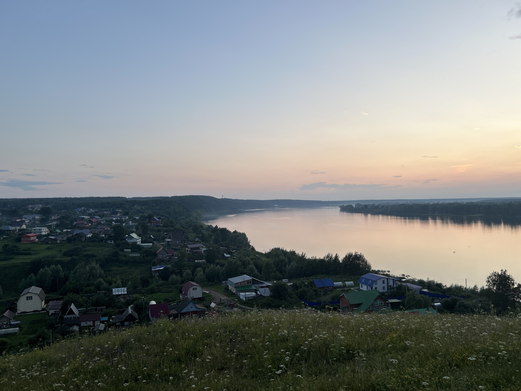 The image depicts a panoramic view of a rural settlement along a wide river, likely at dusk. The foreground is a grassy hillside dotted with small white flowers. Beyond the hill lies a cluster of houses with various roof colors, predominantly red, green, and blue, nestled among trees and vegetation. A road appears to wind through the settlement. The river dominates the middle ground and extends into the distance, reflecting the soft hues of the twilight sky. The sky transitions from a pale blue at the top to a warm peach and pink near the horizon, with a few faint clouds visible. The overall impression is one of a serene, tranquil evening in a village setting. There are no people visible in the image. The location is identified as Olkhovka, Russia.