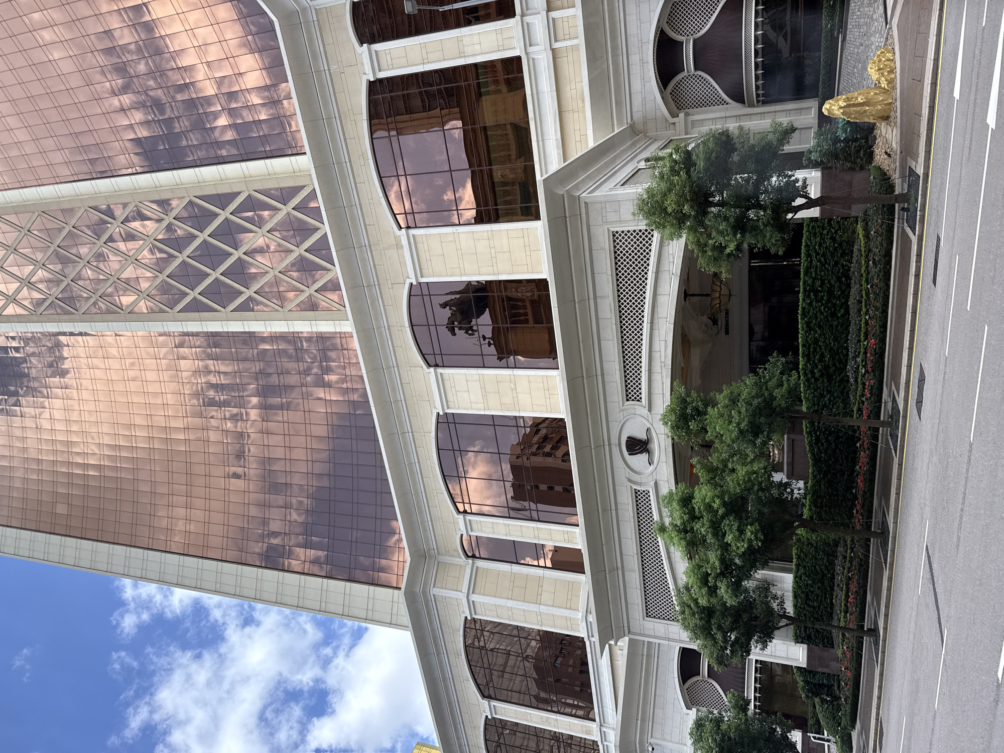 This is a low-angle, wide exterior shot of a large, modern building in Macau, China. The building features a pale, cream-colored stone facade with white trim and large, arched windows with dark, reflective glass. The upper section of the building is clad in rose-gold glass, reflecting the sky and clouds. The foreground shows a paved street with white lane markings and a yellow curb. A sidewalk with several trees, hedges, and flower beds separates the street from the building's entrance. The building's entrance is grand, with a large, arched overhang supported by columns. A gold-colored rock sculpture is visible to the right of the entrance. The sky is blue with scattered white clouds.