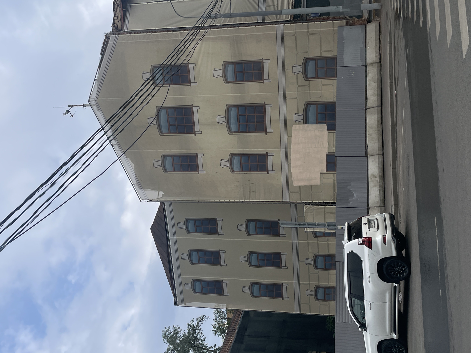 The image depicts a street scene in Moscow, Russia, featuring a large building undergoing renovation. The building is covered in a large tarp that has been printed with a façade of windows and architectural details, creating an illusion of a finished structure. The tarp is secured with ropes and wires, and a weather vane is visible on the roof. In front of the building, there is a construction fence made of concrete blocks and metal sheeting. To the left, a white Lexus GX SUV is parked on the street, facing away from the camera. The street is paved, and a crosswalk is visible in the foreground. The sky is overcast with scattered clouds. The overall impression is of a city undergoing development and restoration.