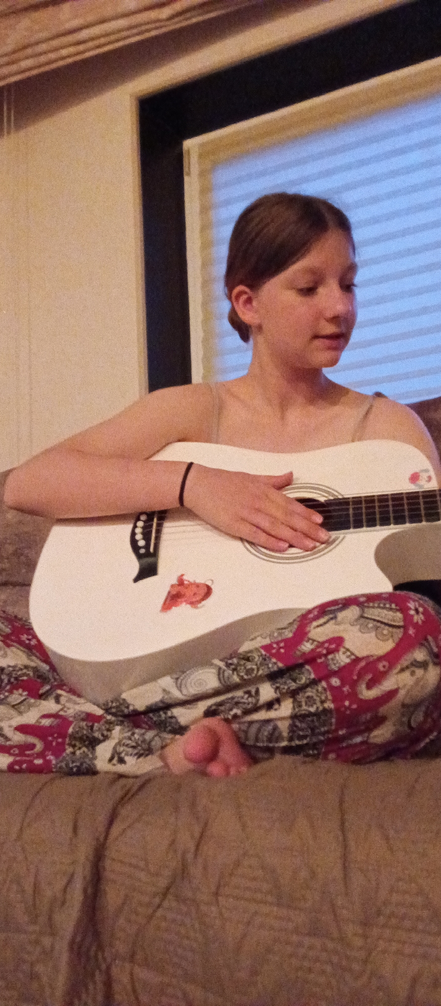 A young Caucasian female with light brown hair pulled back in a bun is seated on a brown upholstered couch and playing a white acoustic guitar. She is wearing patterned pajama pants with a red and white elephant design. A black elastic bracelet is on her left wrist. A sticker of a red cartoon character is on the body of the guitar. The background includes a white wall with a brown trim and a window with closed blinds. The location appears to be indoors, possibly a residence. City and location is unknown.