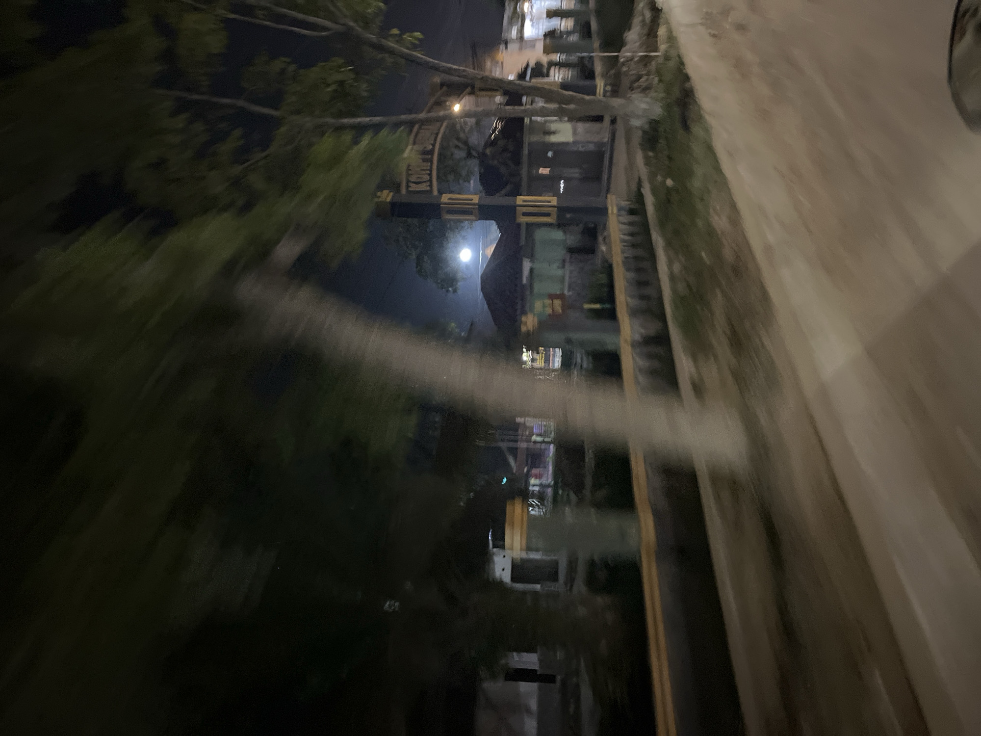 The image depicts a nighttime scene in Bundar, Indonesia, likely taken from a moving vehicle. The perspective is tilted, creating a sense of motion blur, especially in the foreground where horizontal streaks of light are visible.
The scene features a street with buildings and trees lining one side. There's a prominent gate or archway with black and yellow markings, possibly indicating an entrance or a boundary. A bright light, possibly the moon, is visible in the sky, illuminating the area. The buildings appear to be commercial establishments or residences, with some windows showing lights. Trees with dark foliage are scattered throughout the scene. The ground beside the road seems to be a dirt or unpaved area, with some vegetation.
The overall atmosphere is dark and somewhat blurry due to the low light conditions and movement. The image captures a slice of everyday life at night in a town or village setting. There is visible text on the archway, but it is largely illegible due to the blur and resolution. The image does not suggest any specific activity or event, but rather a general view of a street at night.