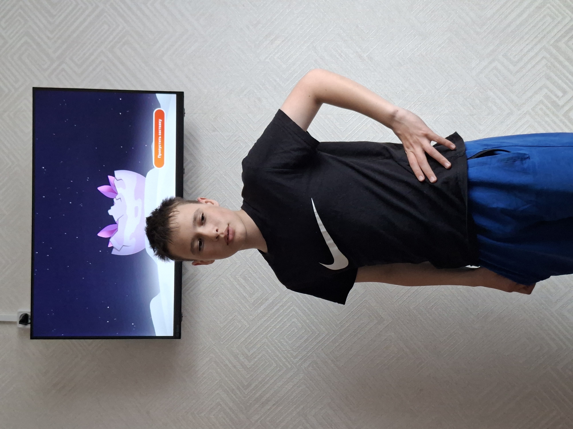 The image features a young person, likely a boy, standing in an indoor setting. He is wearing a black t-shirt with a white Nike swoosh logo and blue pants. His left hand is on his hip, and he is looking directly at the camera with a neutral expression.
Behind him, the wall is covered with light-colored wallpaper that has a repeating geometric pattern of interconnected squares and lines. To his left, a flat-screen television is mounted on the wall. An electrical outlet and a white cable are visible beneath the TV.
The television screen displays a vibrant animated scene or a video game interface. The background is a dark blue, resembling a starry night sky with many small white dots. Large, cloud-like formations, predominantly white, are also visible. A distinct object or character with intricate designs in shades of purple and pink is featured on the screen. In the upper right corner of the TV display, there is an orange rectangular button or banner with white text that reads "Пропустить заставку," which is Russian for "Skip intro."
The image itself is rotated 90 degrees counter-clockwise, making the boy and the television appear sideways. The lighting is uniform, suggesting an indoor light source.