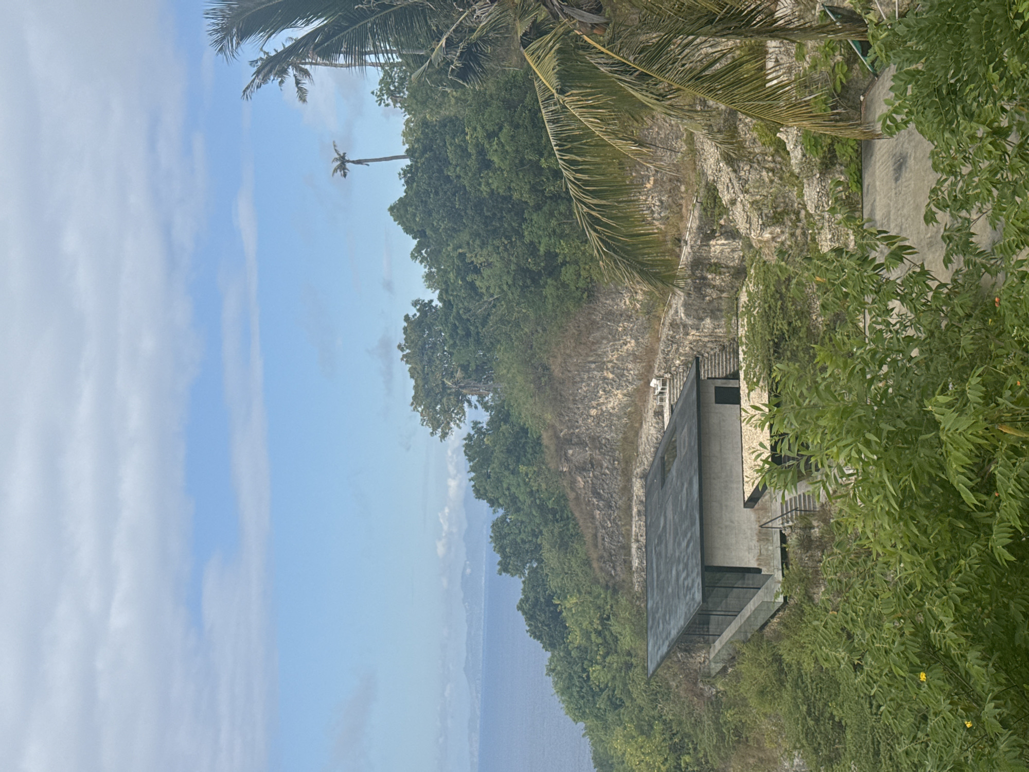 The image depicts a modern villa nestled into a lush, tropical hillside overlooking the ocean. The villa features a sleek, dark-tiled roof and extensive glass walls that offer panoramic views. A concrete structure with a stepped entrance leads to the main living area. The surrounding landscape is characterized by steep, rocky inclines covered in dense green foliage and palm trees, typical of the island's terrain. In the distance, the calm blue sea stretches towards a hazy horizon, with faint outlines of other islands visible. The sky is partly cloudy with patches of blue. The scene appears serene and secluded, embodying a luxurious retreat in a natural setting. The location is identified as Nusa Penida, Indonesia, a popular island destination known for its dramatic cliffs and pristine waters. There are no people visible in the image.