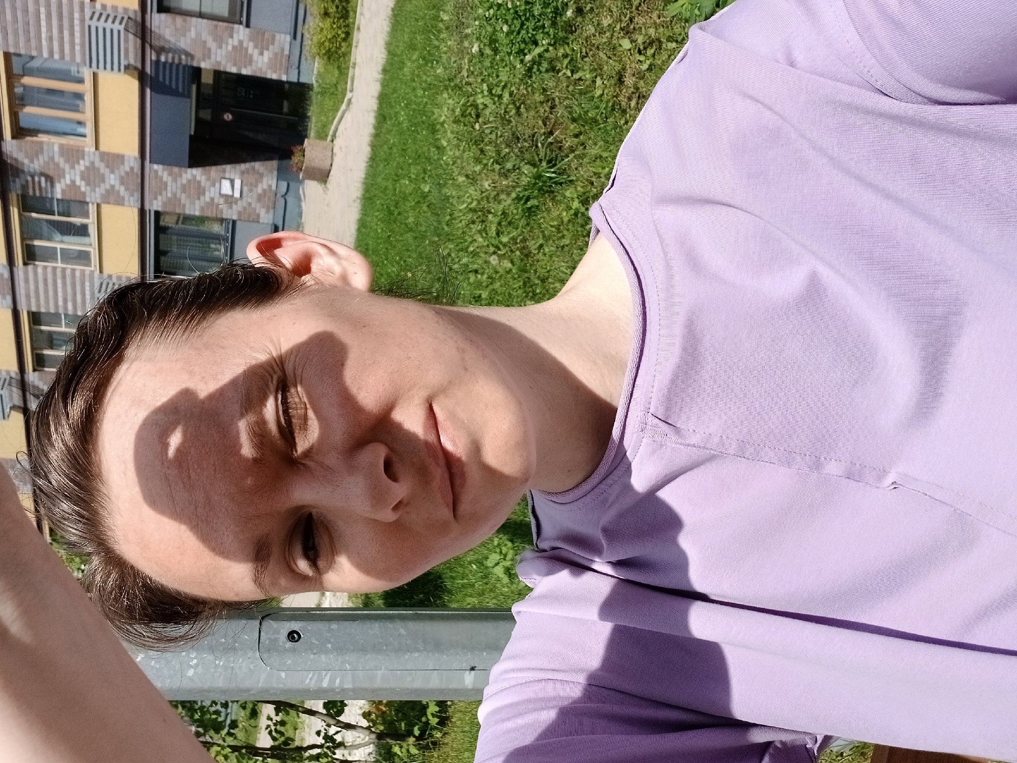 A mid-shot, eye-level selfie of a woman taken outdoors on a sunny day. The woman is positioned in the center of the frame, looking slightly to her left. She has light skin, short brown hair, and is wearing a light purple t-shirt. Her left eye is squinting against the sun, and her right arm is raised, casting a shadow over her forehead. She appears to be standing in a grassy area with a paved walkway in the background. Behind the walkway is a modern, multi-story building with yellow and brick-patterned facade, and apartment windows. The location is Yekaterinburg, Russia.