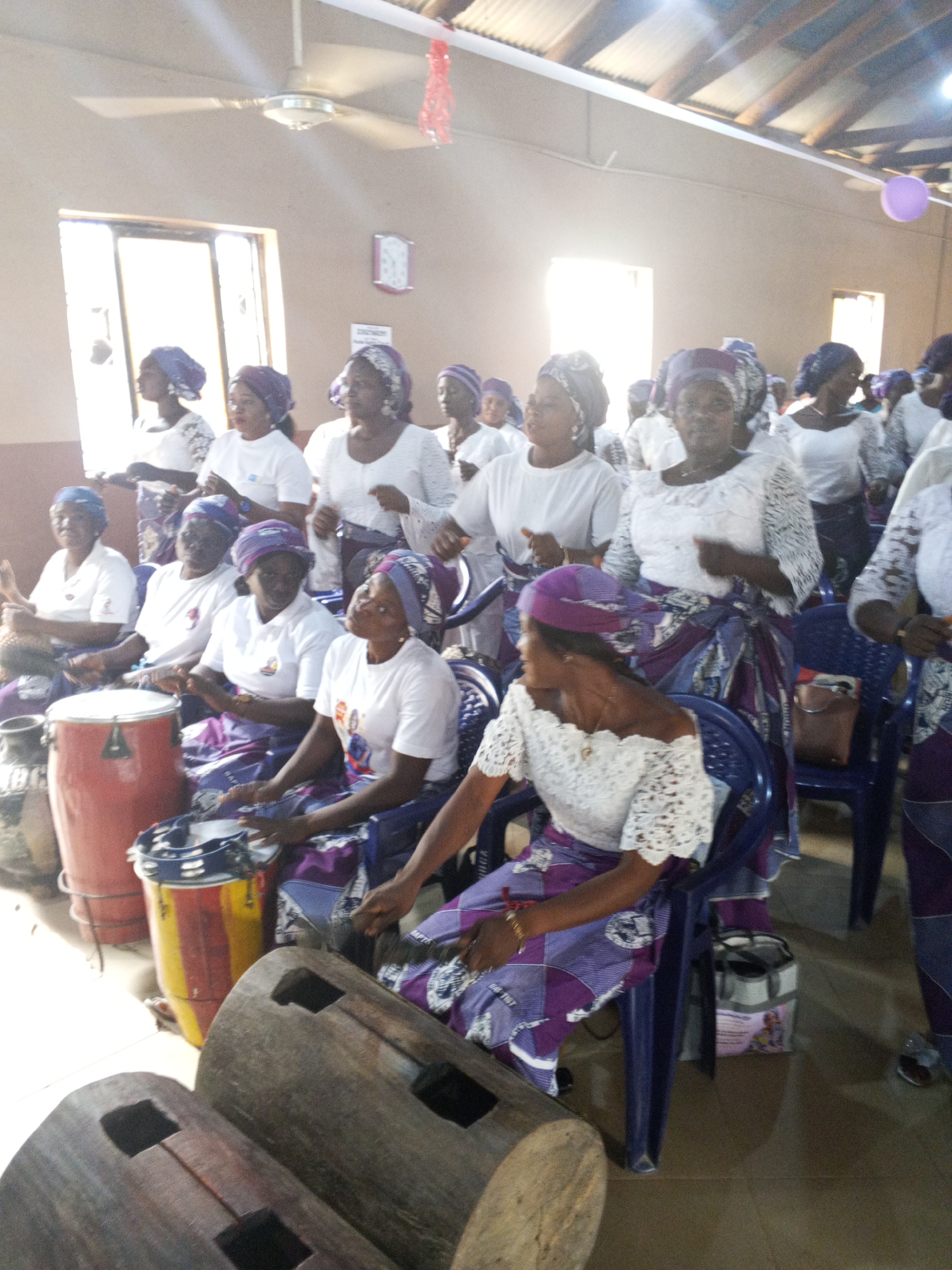 Inside an indoor location in the city of Unknown, Unknown, a group of adult women, predominantly black, are engaged in a musical performance or religious activity. Most of the women are wearing white tops or dresses paired with purple headscarves and purple and white patterned skirts. Some women are standing and singing, while others are seated and playing musical instruments. Instruments include traditional wooden slit drums, tamborines, and a tall cylindrical drum. The scene is illuminated by natural light streaming in through windows, with an additional ceiling fan visible. A purple balloon hangs in the upper right corner of the frame.