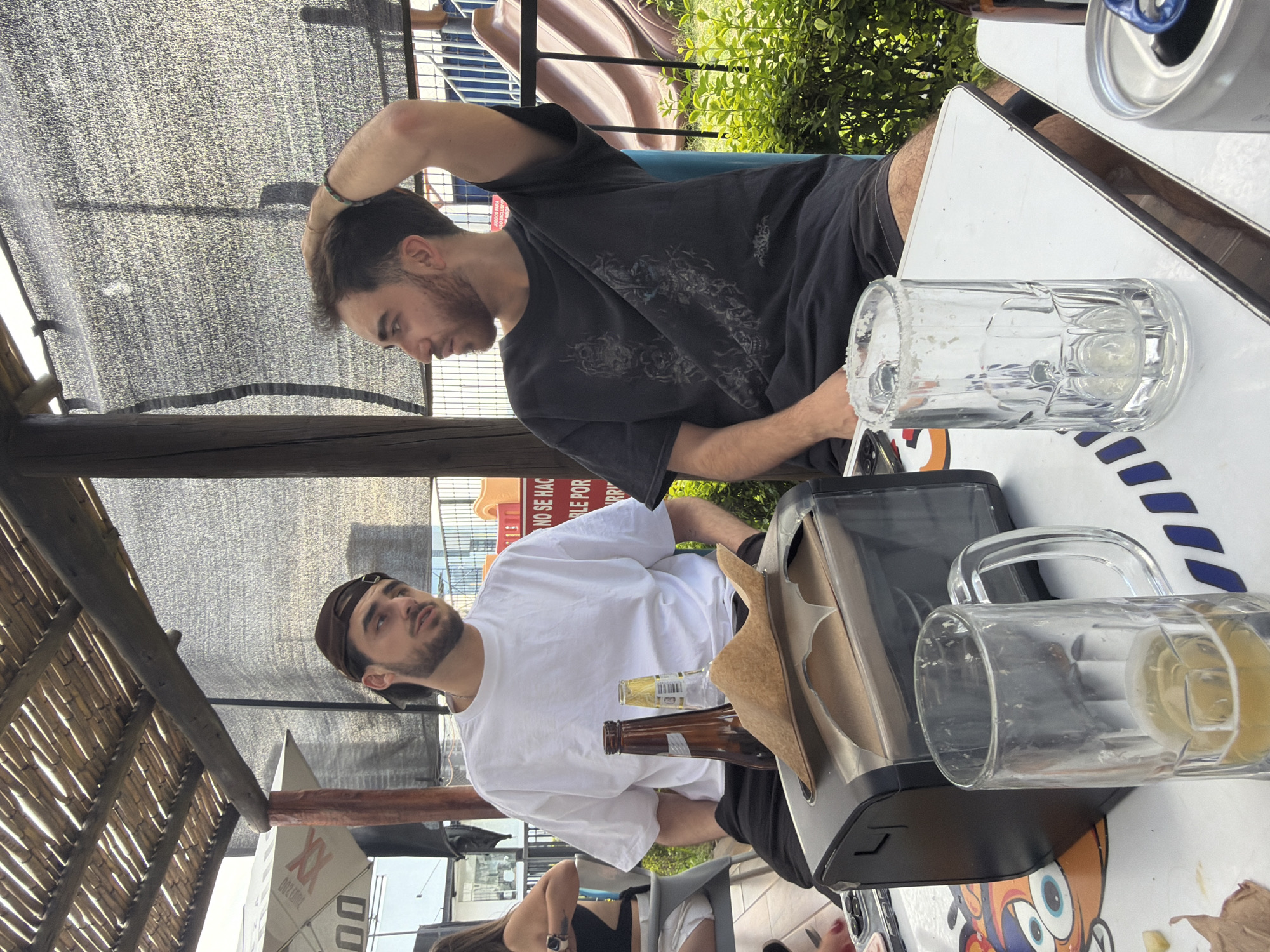 The image depicts two men seated at an outdoor table, likely at a restaurant or bar. One man, wearing a dark t-shirt, is touching his head, while the other man, wearing a white shirt and a cap, is looking forward. The table holds a tray, empty beer bottles, a beer mug, and a can of beer. A third person is partially visible sitting at the table. The setting appears to be San Bernardino Tlaxcalancingo, Mexico, based on the table and the signs near the two men. The location is an outdoor venue, possibly a restaurant or bar with a thatched roof structure.