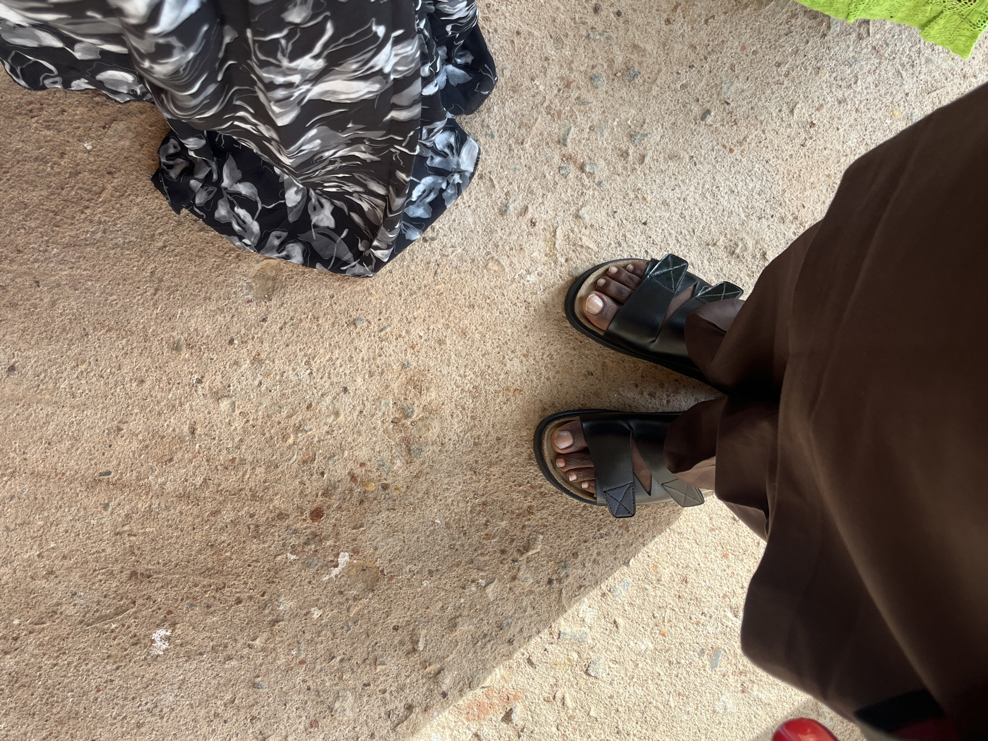 The image shows a top-down view of a person's feet wearing black sandals on a rough, sandy-colored ground. A dark brown garment is draped over the person's legs. To the right, a patterned fabric with black and white swirls is visible, and to the far right, a neon green textured fabric is present. A shadow falls diagonally across the ground from the left. The scene is set outdoors in Bukuru, Nigeria.
