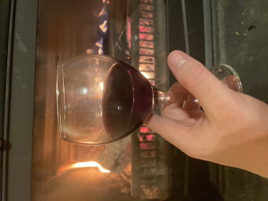 The image depicts a human hand holding a wine glass containing dark red liquid in front of an active fireplace, set in Palež, Montenegro. The hand, exhibiting light-toned skin, is positioned in the lower-center of the frame, grasping the stem of the clear glass. The glass holds an estimated 80-100 ml of liquid. The background is dominated by the interior of a fireplace or wood-burning stove, where orange and blue flames are visible consuming logs. Red-hot embers glow at the base of the fire, resting on a dark-colored grate. Reflections from the fire are visible on the surface of the wine and the glass itself. The scene is illuminated primarily by the firelight.