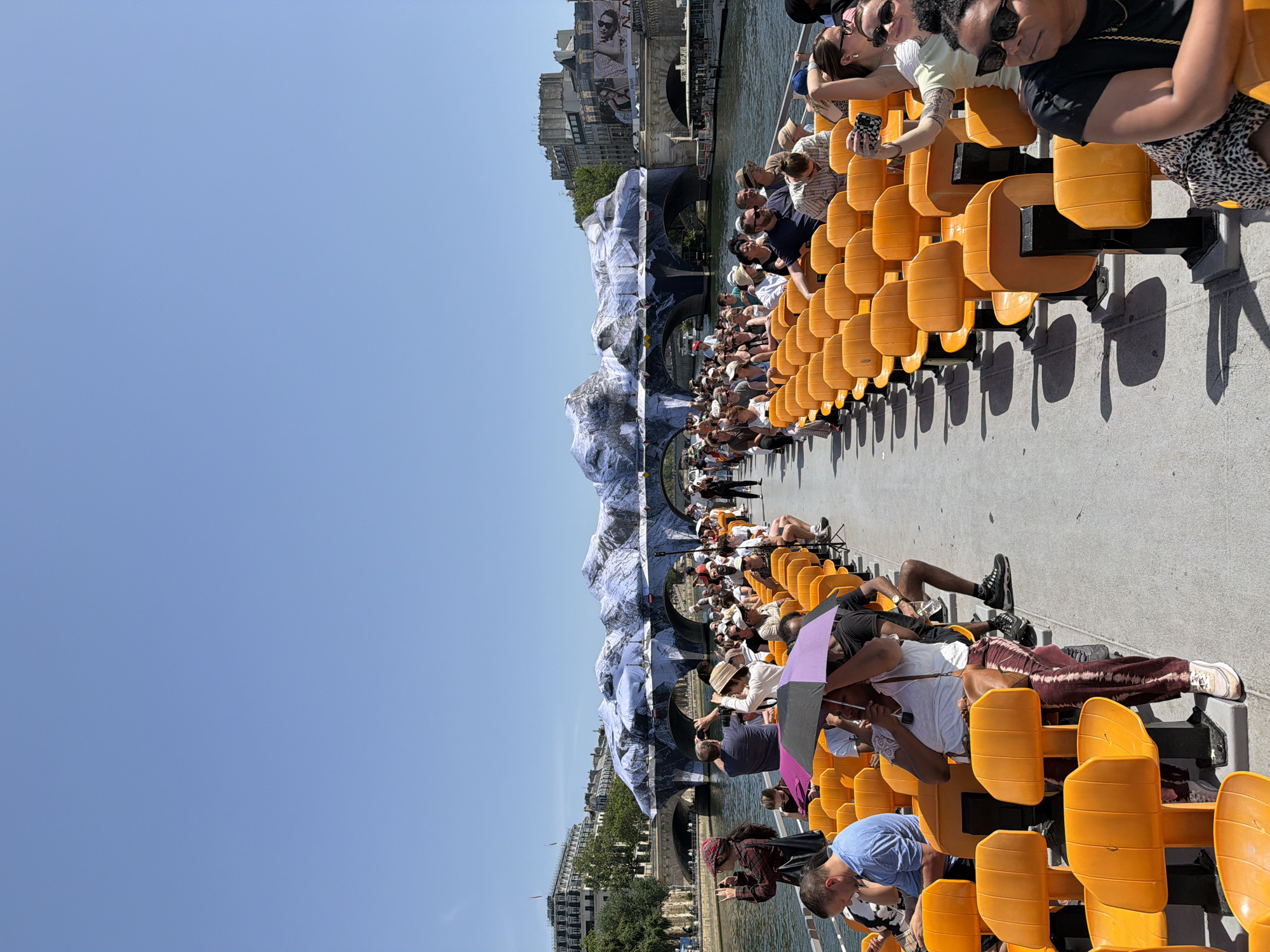 People are seated in rows of orange stadium-style seats, likely on a boat, facing a building or bridge decorated with what appears to be a mountain scene, possibly painted or projected. The location appears to be on the Seine River in Paris, France, judging by the architecture of the buildings. The clear blue sky suggests a sunny day. People are taking pictures and generally observing the scenery.