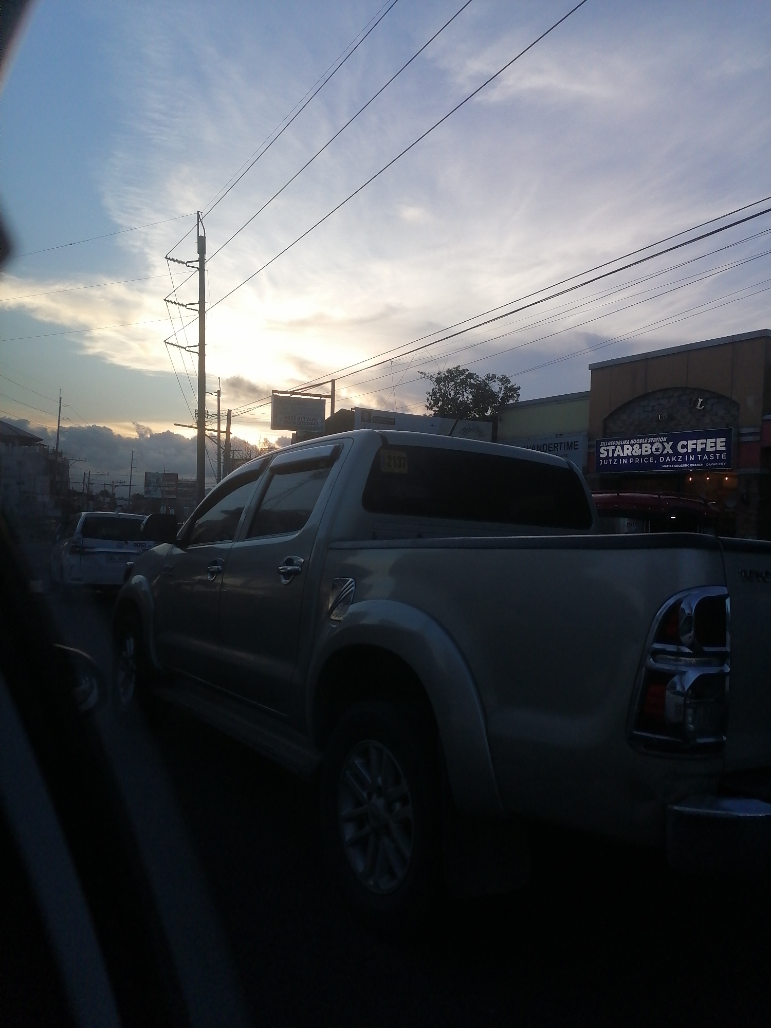 The image depicts a street scene at dusk in Matina Crossing, Davao City, Philippines. The sky transitions from light blue at the top left to a bright yellow-orange near the horizon on the right, indicative of sunset, with scattered cirrus and stratus clouds present. Numerous utility poles and a dense network of overhead power lines crisscross the sky and midground.
In the immediate foreground, the rear three-quarter view of a light-colored Toyota Hilux pickup truck is prominent, appearing stationary. Its rear wheels, tailgate, and taillight assembly are visible. Partially obscured behind it, a white pickup truck with a visible license plate segment reading "4061" is also stationary.
On the right side of the midground, a commercial building with an orange-brown facade and a stone-textured section houses "STAR&BOX COFFEE" and "ZILI REPUBLIKA NOODLE STATION." The sign explicitly states "MATINA CROSSING BRANCH - DAVAO CITY." To its left, an adjacent building with a lighter yellow facade shows a partial sign with the word "WANDERTIME." Further left, silhouetted buildings and trees line the street. No discernible people are visible. The scene is illuminated by the fading natural light of sunset, casting soft shadows across the vehicles and structures.
