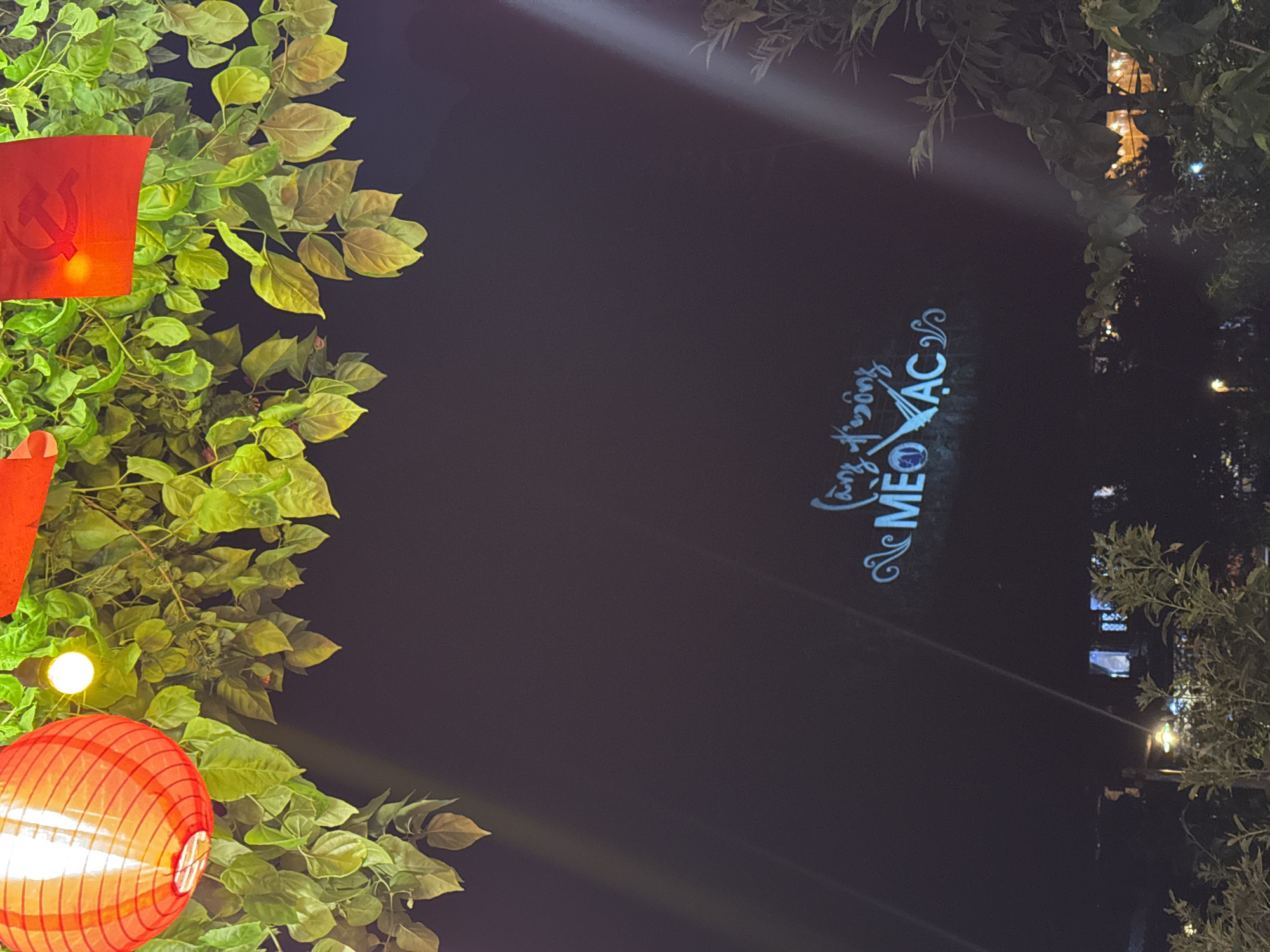 The scene captures an outdoor nighttime ambiance in the Mèo Vạc Commune, Vietnam. The left of the frame is filled with lush foliage, intertwined with a red communist flag and a traditional red lantern emitting a warm glow. The central part of the image transitions to a darker space, illuminated by a faint beam of light, which seems to project a light-blue illuminated script “Mèo Vạc”. On the right side, there is more foliage, and faint distant structures suggesting the presence of buildings.