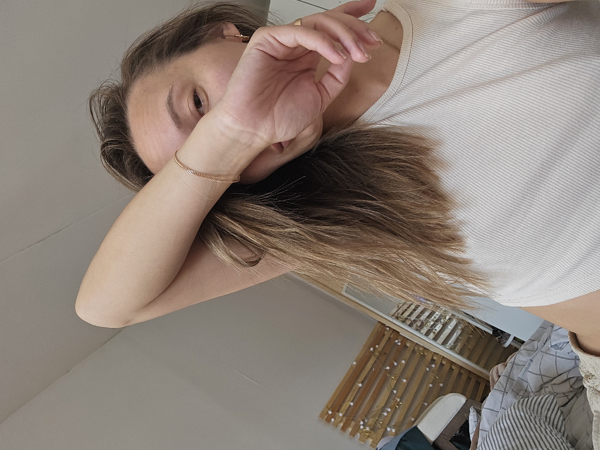 A light-skinned person with long, light-brown hair is lying down in a bedroom. They are wearing a light-colored ribbed tank top and have a gold bracelet on their left wrist. Their left hand is partially covering their face. The room has a window with a wooden shade, bedding with a checkered pattern, and a white shoe visible on the bed. The location is not discernable.
