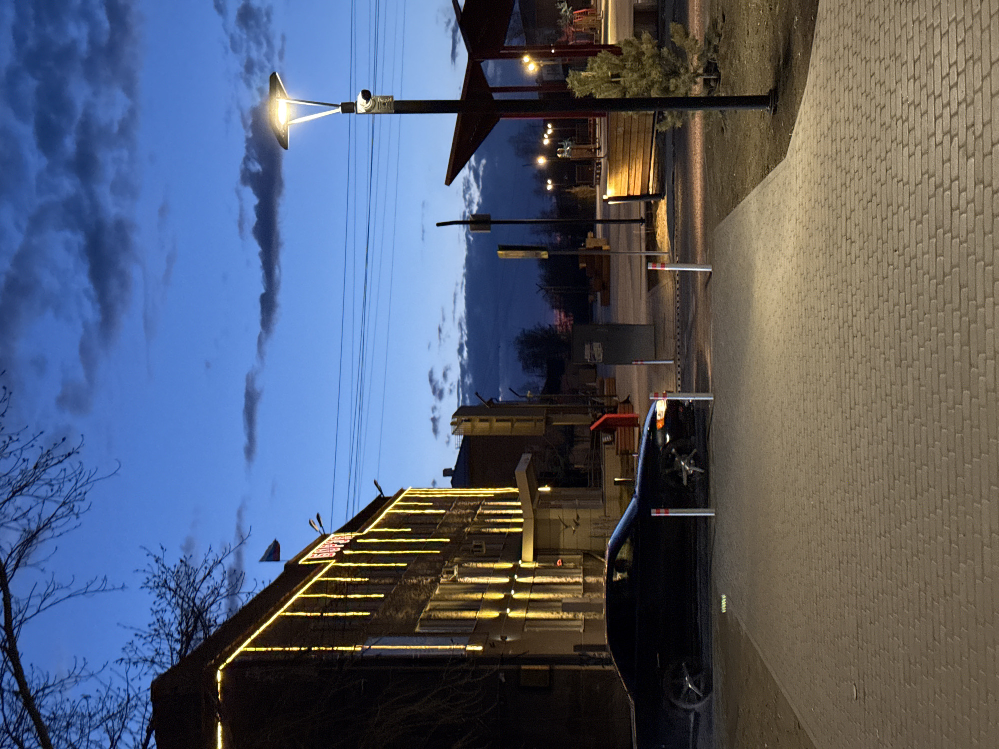 Captured at dusk in Borzya, Russia, the image showcases a street scene. A paved walkway extends along the right side of the frame. A dark-colored car is parked adjacent to the walkway. To the left, a building is illuminated with decorative yellow lights tracing its roofline. In the background, street lamps cast a glow on the area, and utility poles are visible against the darkening sky with scattered clouds. The overall atmosphere suggests a quiet evening.