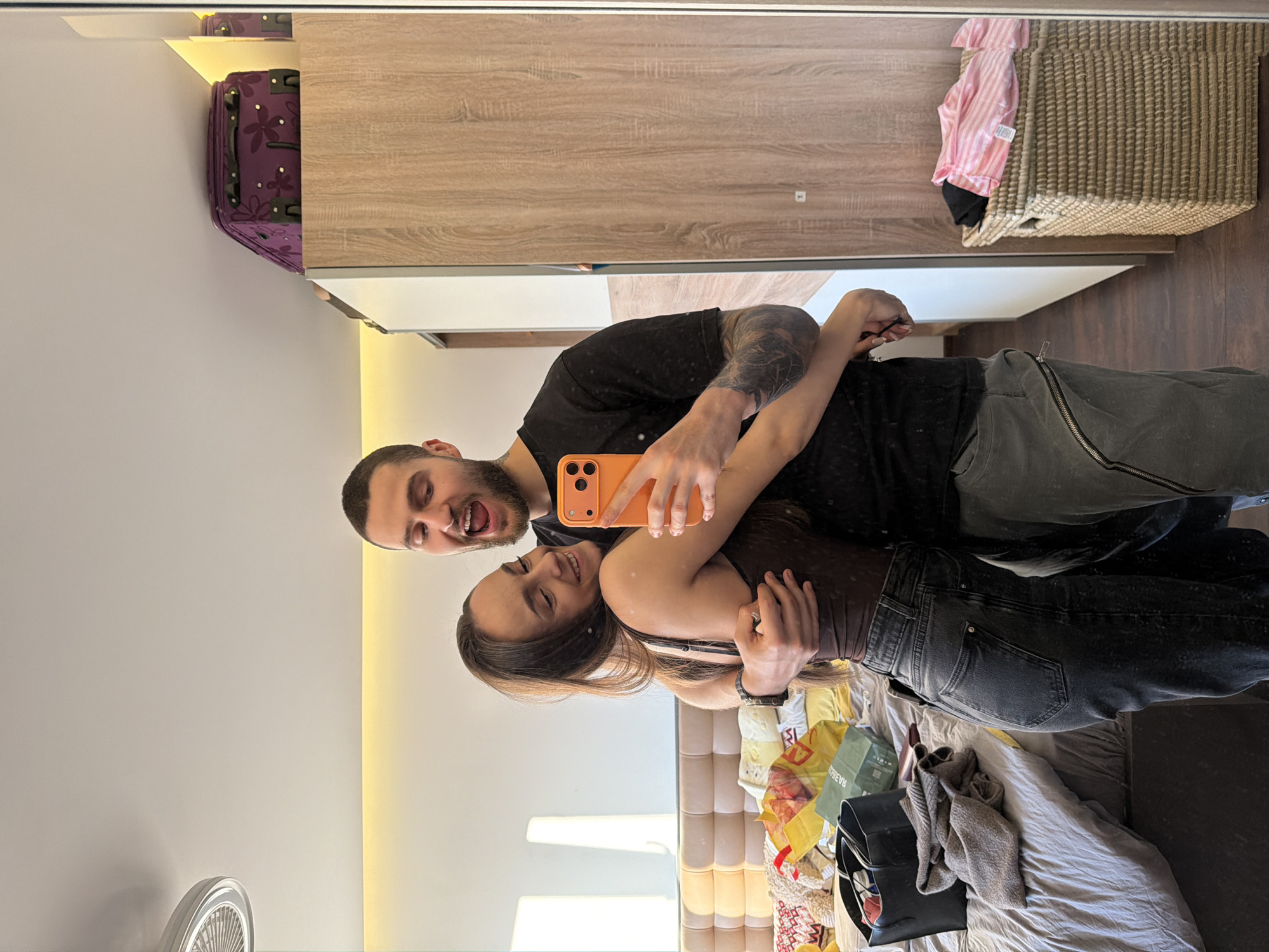 A selfie in a room, likely in Sofia, Bulgaria, features a man and a woman embracing. The man, with a beard and tattoos on his arm, holds an orange phone. The woman has long hair and is smiling. They are in front of a mirror, capturing a cluttered room. A bed with scattered items, including shopping bags and clothes, is visible. In the background, there is a wooden wardrobe, a suitcase, and a wicker basket.