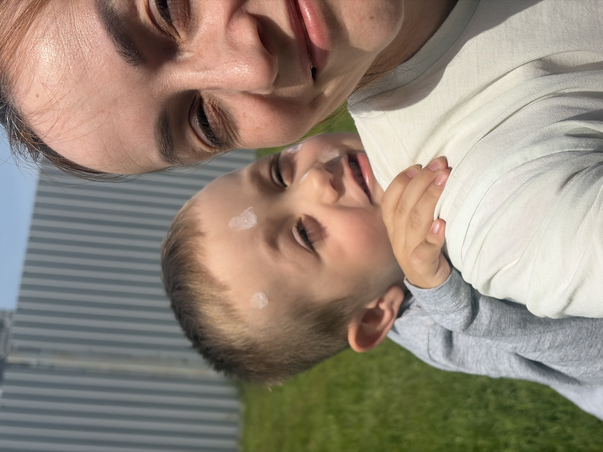 A selfie shows a woman with reddish-brown hair holding a young child in her arms. Both are looking at the camera. The child is wearing a gray long-sleeve shirt. The woman is wearing a white shirt. There are two spots of white cream on the child's forehead. The background shows a corrugated metal facade and grass. Based on the geolocation information, the photo was taken in Chaikovsky, Russia.