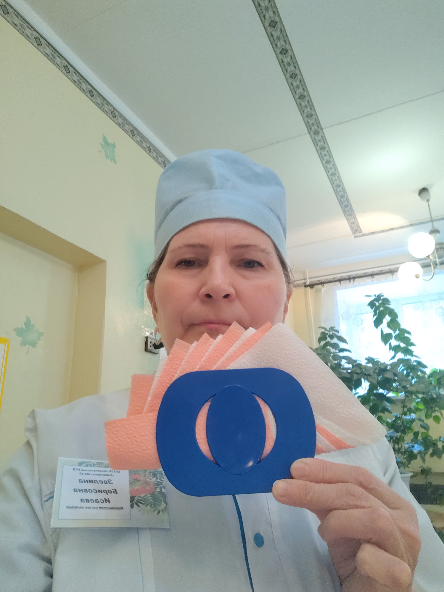 A woman in a blue surgical cap and white lab coat holds a blue eye chart ring with peach-colored tissue paper behind it. She looks directly at the camera with a neutral expression. A name tag is visible on her left side. In the background, there is a wall with a light yellow hue and a light fixture hanging from the ceiling. A green leafy plant is near a window. The name tag says "Nozdracheva Valentina Viktorovna Medical Sister Of The Dispensary Sarapul, Russia". The photo was likely taken in Sarapul, Russia.