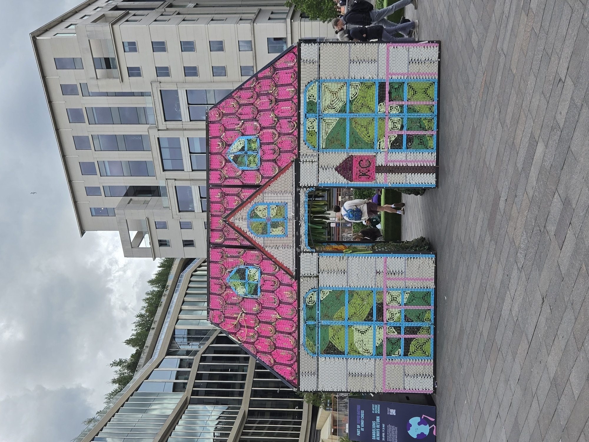 Here's a detailed description of the image:
The image showcases a colorful, whimsical art installation depicting a house, situated in what appears to be a public space, possibly in the city of London, United Kingdom. The structure is made of what looks like woven or interlaced materials, with a pink roof designed with geometric shapes and light-colored walls adorned with depictions of windows. The windows have a green, blue and turquoise design. The house has a very small KC label that has a pink background.
Several buildings are visible in the background. One is a modern, multi-story building with many windows. Another building, visible to the left, features a unique, curved facade with reflective windows and green vegetation.
People are present in the scene, some appearing to be passersby and others possibly interacting with the artwork. There are people walking past and others looking inside.
On the floor, directly in front of the house installation, there is a small dark blue sign, it includes the words: PART OF SUPER NATURE AT KINGS CROSS, ARTIST DELPHINE DÉNÉRÉAZ.
The ground is paved with light grey tiles. The sky is overcast with cloudy skies. The image is oriented on its side, with the top of the buildings toward the left side of the image.